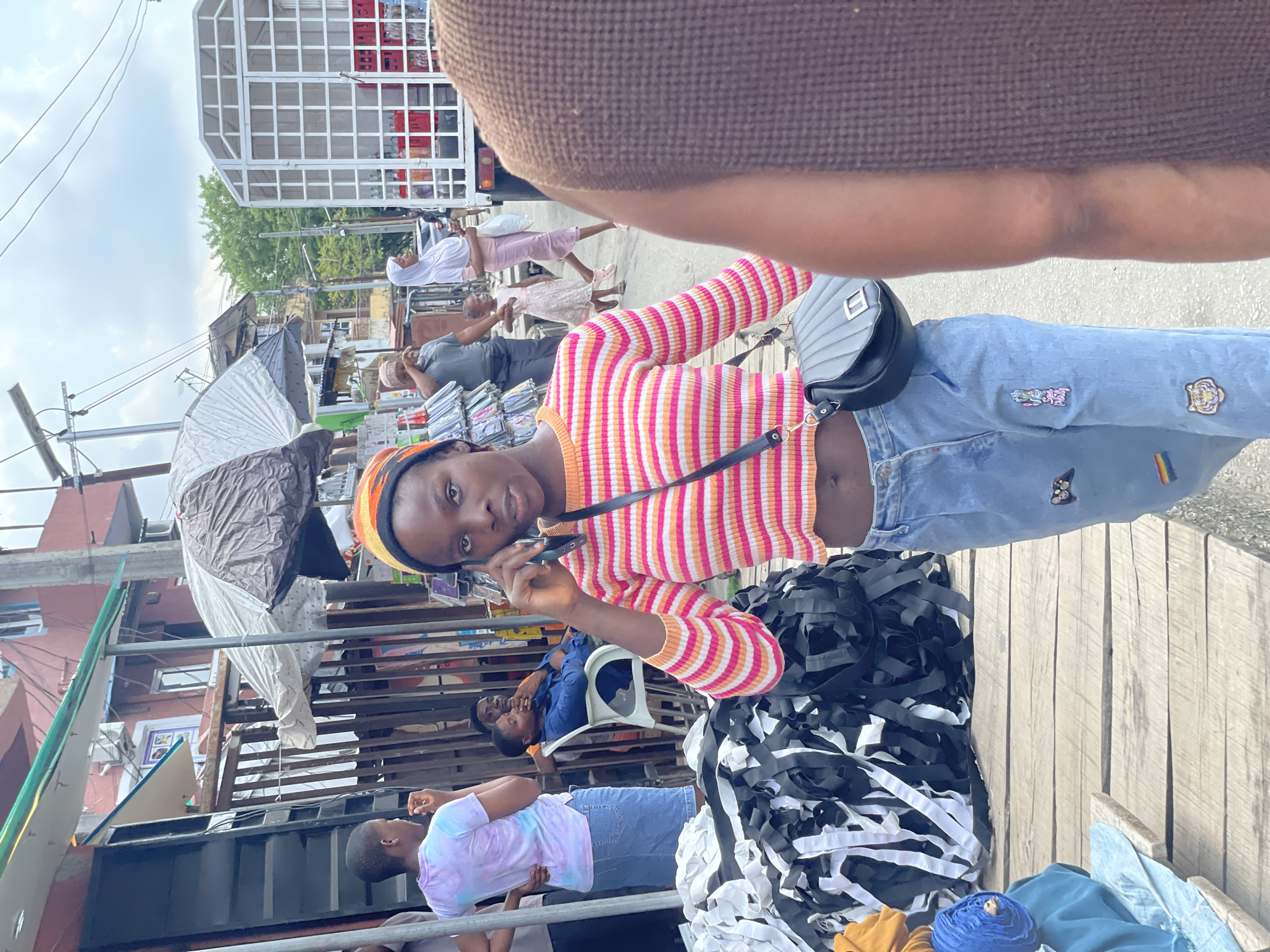 The image captures a vibrant, bustling outdoor street market scene, likely in Lagos, Nigeria, under an overcast sky during daytime.
In the immediate foreground, a young person, possibly female, is the central subject, looking directly at the camera while holding a mobile phone to their ear. They are dressed in a colorful horizontal-striped crop top (pink, orange, yellow, white) and light blue jeans adorned with decorative patches, including a rainbow flag and a butterfly. A black or dark grey shoulder bag hangs across their body. Part of another person's arm and shoulder, wearing a brown textured garment, is visible in the upper right, suggesting the photographer's proximity or another person standing very close.
The foreground also features market goods displayed on a wooden surface, including large piles of black and white fabric strips or ribbons, and rolls of blue and yellow textiles. To the main subject's left, another individual in a light blue/purple tie-dye t-shirt and jeans stands looking away.
The mid-ground and background reveal more of the market activity. Several people are visible, including two individuals seated under a large grey umbrella, and others walking further down the street, one wearing a white head covering and a light pink outfit. Market stalls, makeshift shelters, metal structures, and residential buildings line the street. A white truck with a distinctive caged cargo area is visible in the distant background. Overhead power lines crisscross the sky. No clearly legible text is visible on signs or clothing.