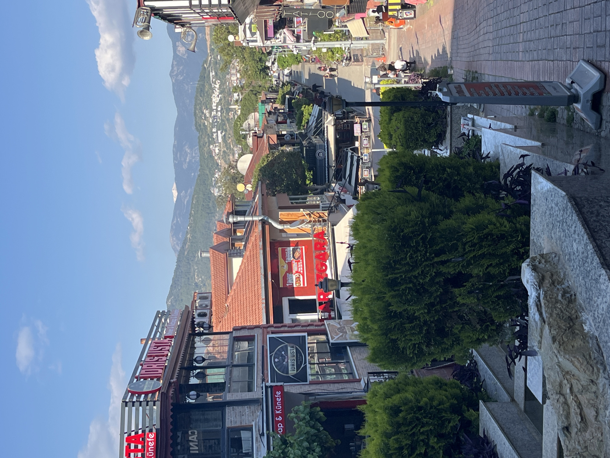 The image captures a vibrant, sunny daytime street scene in Alanya, Turkey, with mountains forming a dramatic backdrop. The perspective is slightly elevated, looking down a bustling commercial street lined with multi-story buildings that house various shops and restaurants.
The clear blue sky, dotted with fluffy white clouds, indicates fair weather. A prominent mountain range rises in the distance, its lower slopes speckled with white buildings. The street below is paved with light-colored bricks, and people can be seen walking and sitting at outdoor tables, enjoying the cafes and restaurants.
Numerous commercial signs are visible:
* A large red sign on a building reads "ADAG...STA" (partially obscured, likely "ADAGUSTA").
* Below it, a smaller sign displays "TA..." and "Çiğ köfte & Künefe".
* A black sign further down the street says "İDRİS USTA".
* In the distance, a "TATTOO" sign is visible, along with another red "AGA" sign and a "PASTA" sign.
* A reflected word "CANI" can be seen on a window.
The buildings feature various architectural details, including red-tiled roofs and numerous air conditioning units. Decorative brass musical instruments, including a trumpet and a French horn, are mounted on a building facade near the top left. Green hedges and various plants, some with purple foliage, line the street and are placed in planters, including a textured gray stone retaining wall or planter system on the right side of the frame. A gray and white street sign is also partially visible on the right. The overall impression is a lively Mediterranean town enjoying a bright day.