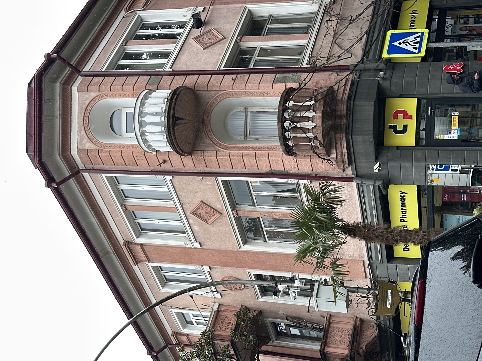 A slightly tilted, upward-angle shot captures a building facade in Batumi, Georgia. The building is characterized by its pinkish-beige color, ornate windows, and a projecting balcony.
On the street level, a 'Dee Zee Pharmacy' sign is visible in yellow. Adjacent to it, a person wearing a dark hooded jacket stands near the entrance of the pharmacy. Above, a yellow sign displays Georgian script alongside a pedestrian crossing sign.
A black car is partially visible in the foreground. A palm tree is partially obscuring the building behind it.