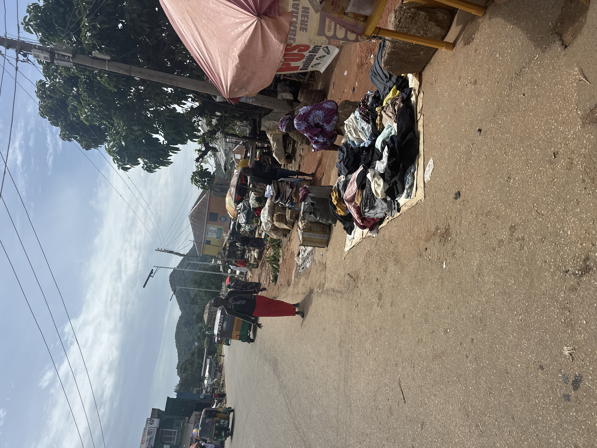 The image captures a street scene in Gwong, Nigeria, likely a marketplace. A woman in a red skirt and black top walks on a paved road. To the right of the road, a stall displays various clothing items spread on the ground and on a mat. A woman in a purple and blue patterned dress sits on a rock near the clothing. A pink umbrella shades part of the stall. Further down the road, a few more people are visible, as are several three-wheeled vehicles (Keke Napeps). Buildings line the street, and in the background, hills are visible under a cloudy sky. The overall impression is of a busy commercial area.