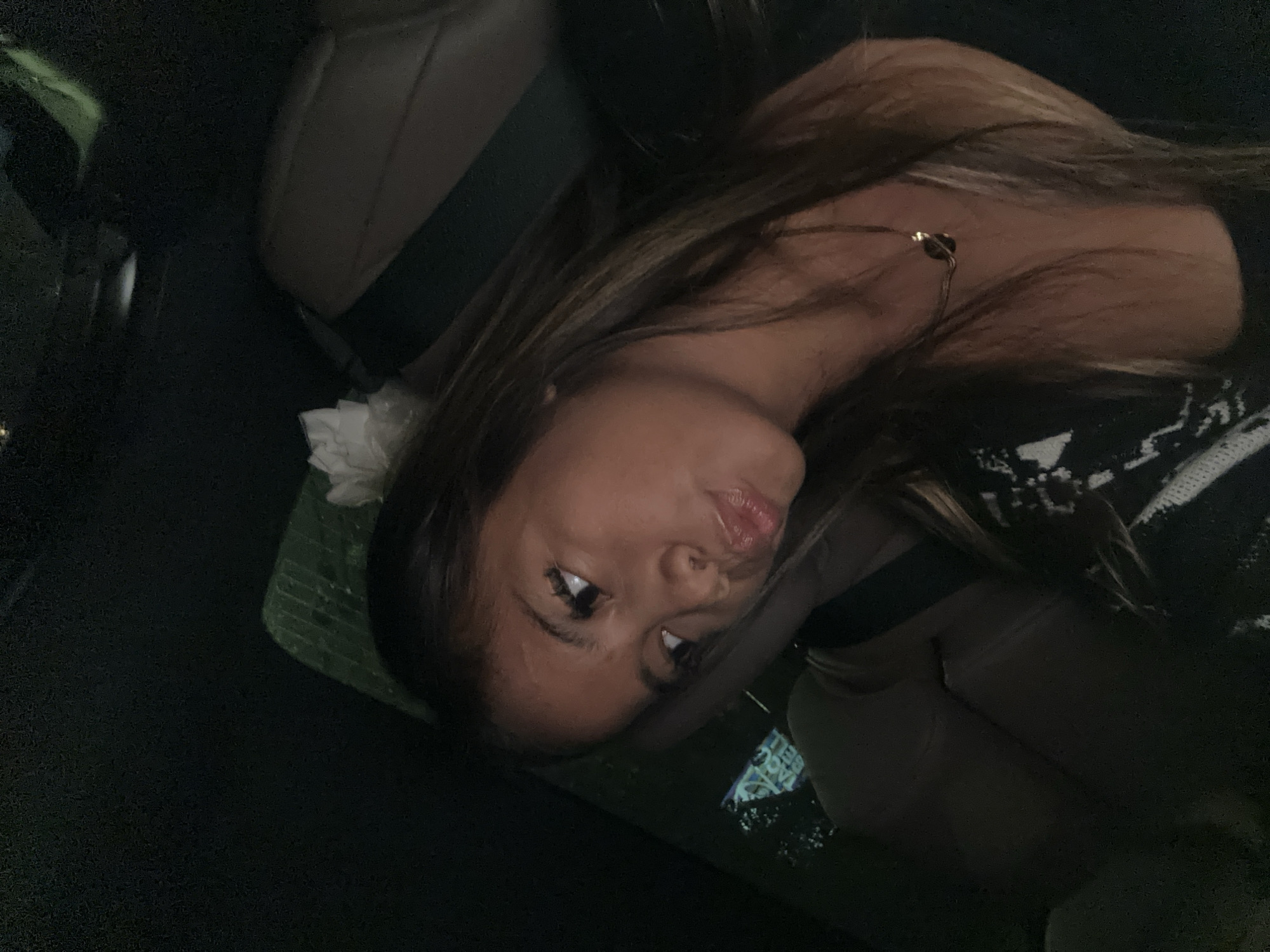 A young woman is reclining in the passenger seat of a car. She has long brown hair with blonde highlights and is wearing a dark top and a gold necklace. A white flower and a green leaf are next to her head. There is a fast-food restaurant logo of "Taco Bell" faintly visible in the background. Based on this information, the location could be South Jakarta, Indonesia, as Taco Bell has locations there. The car's interior is dark, and the seats appear to be beige.