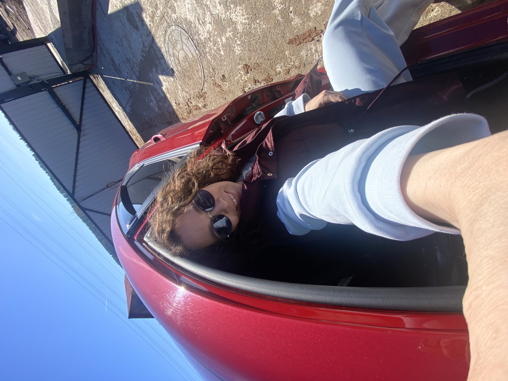 A woman with curly brown hair, wearing sunglasses, a blue long-sleeved shirt, and a maroon vest, is seated in a red car. The car door is open. The location appears to be outdoors, with a building partially visible in the background under a blue sky. No information suggests a specific location like Ольховка, Russia is determinable from the provided image.
