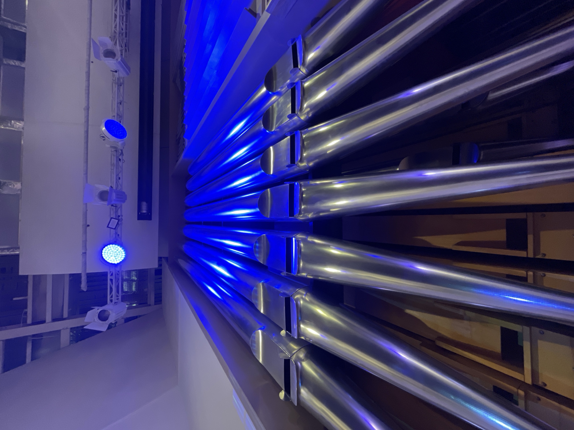 The image presents a close-up, angled view of numerous large, polished metallic pipes, likely part of a pipe organ or a similar architectural installation, brilliantly illuminated by vibrant blue light. The original photograph is oriented vertically, with the pipes appearing to extend diagonally from the bottom-left to the top-right of the frame.
The pipes are silver-colored, highly reflective, and gleam with intense blue highlights and streaks caused by the artificial illumination. Many of the pipes display rectangular openings near their ends, which are characteristic of organ pipes. They are arranged in multiple rows, receding into the background where hints of wooden internal structures are visible.
On the left side of the image, a vertical metal truss structure holds several lighting fixtures. A circular LED spotlight, emitting a powerful blue light, is prominently visible, directing its glow towards the metallic pipes. Above and below this blue LED light, two other white theatrical or architectural spotlights are mounted on the truss.
The setting is an indoor environment, likely a concert hall, auditorium, or a public building in Yekaterinburg, Russia, where such a visually striking display would be installed. No people are present, and no specific activity is taking place. The dramatic blue lighting creates a cool, contemporary, and almost ethereal atmosphere. There is no visible text in the image.