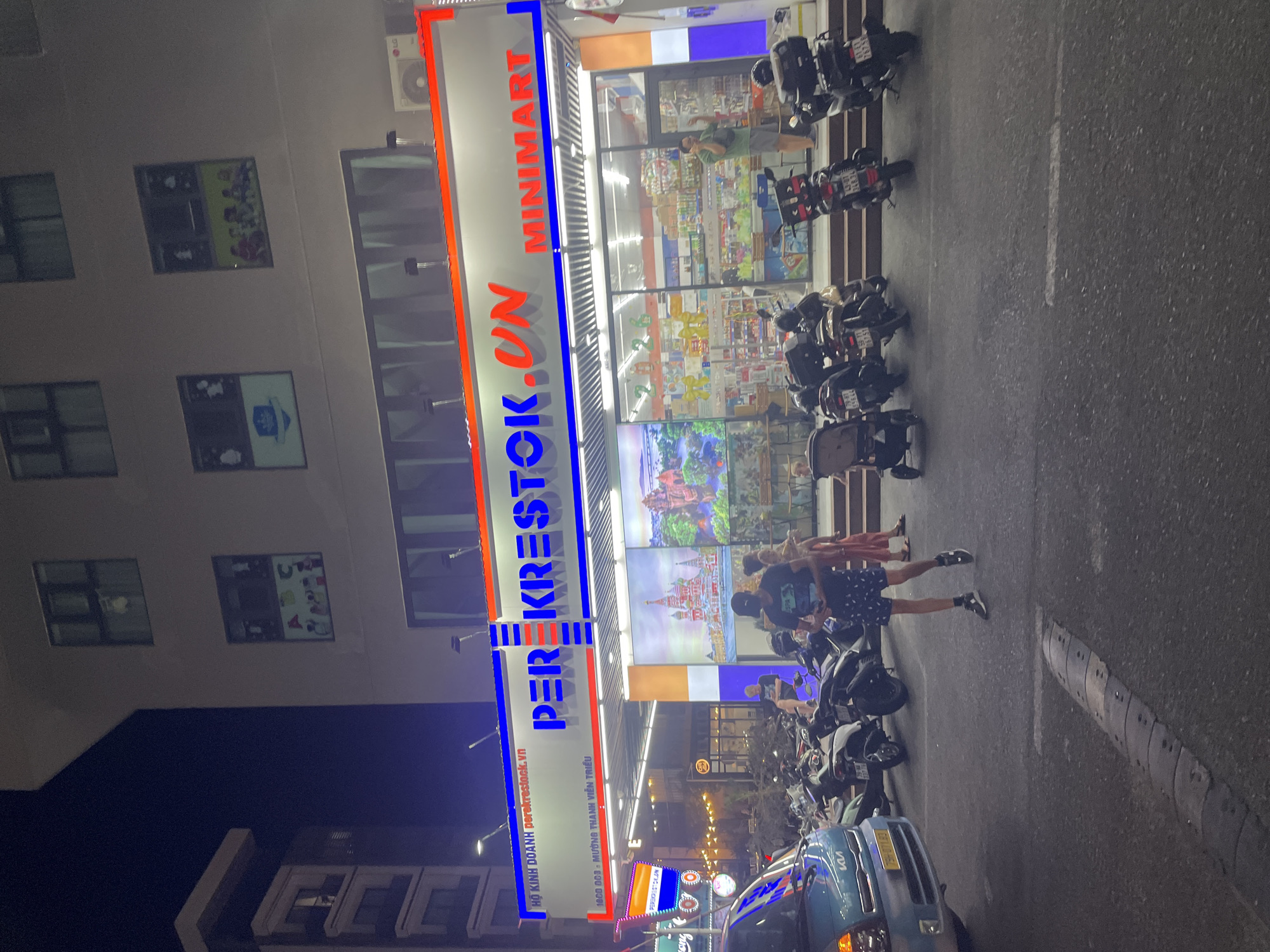 Captured at night in Bắc Nha Trang, Vietnam, a commercial scene unfolds outside a "PEREKRESTOK.UN MINIMART." People are walking along the street, passing by rows of parked motorbikes. The storefront is illuminated, with displays visible through the glass. A blue car is partially visible at the bottom of the frame. Above the minimart, the building's exterior includes windows adorned with decorative images.