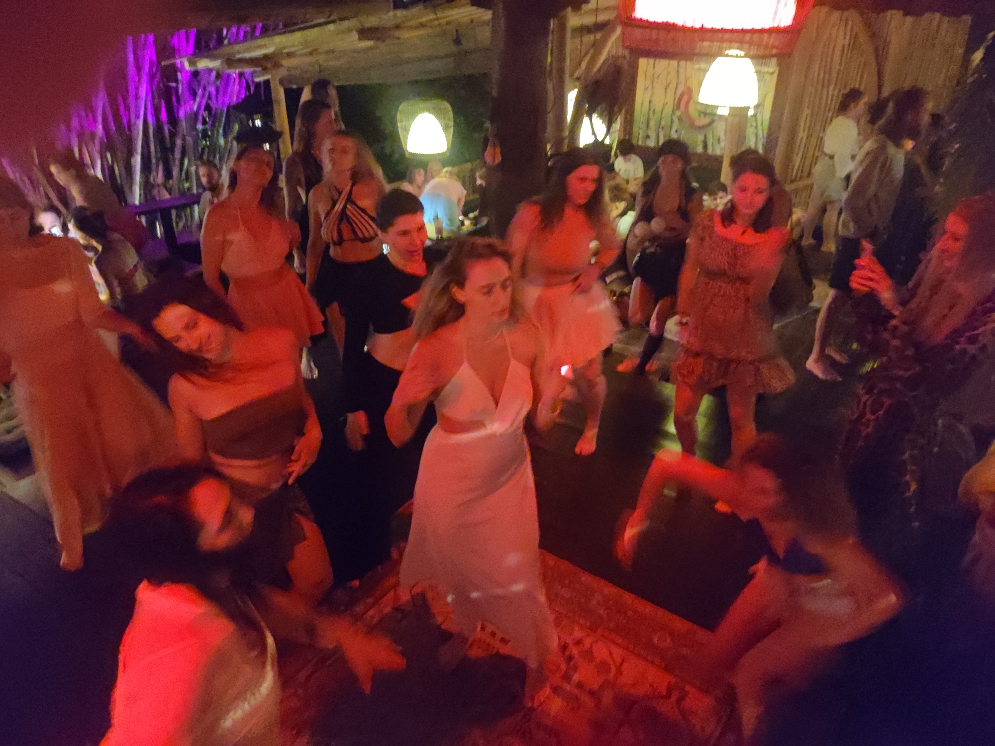 A group of people are dancing inside a building in Lokaserena, Indonesia. The building has a wooden structure and is lit with various colored lights, including purple and red. Most of the people appear to be women, and they are dressed in various casual outfits. They are gathered on a rug and seem to be enjoying themselves. Some people appear to be holding drinks. The lighting is dim, which makes it difficult to discern specific details.