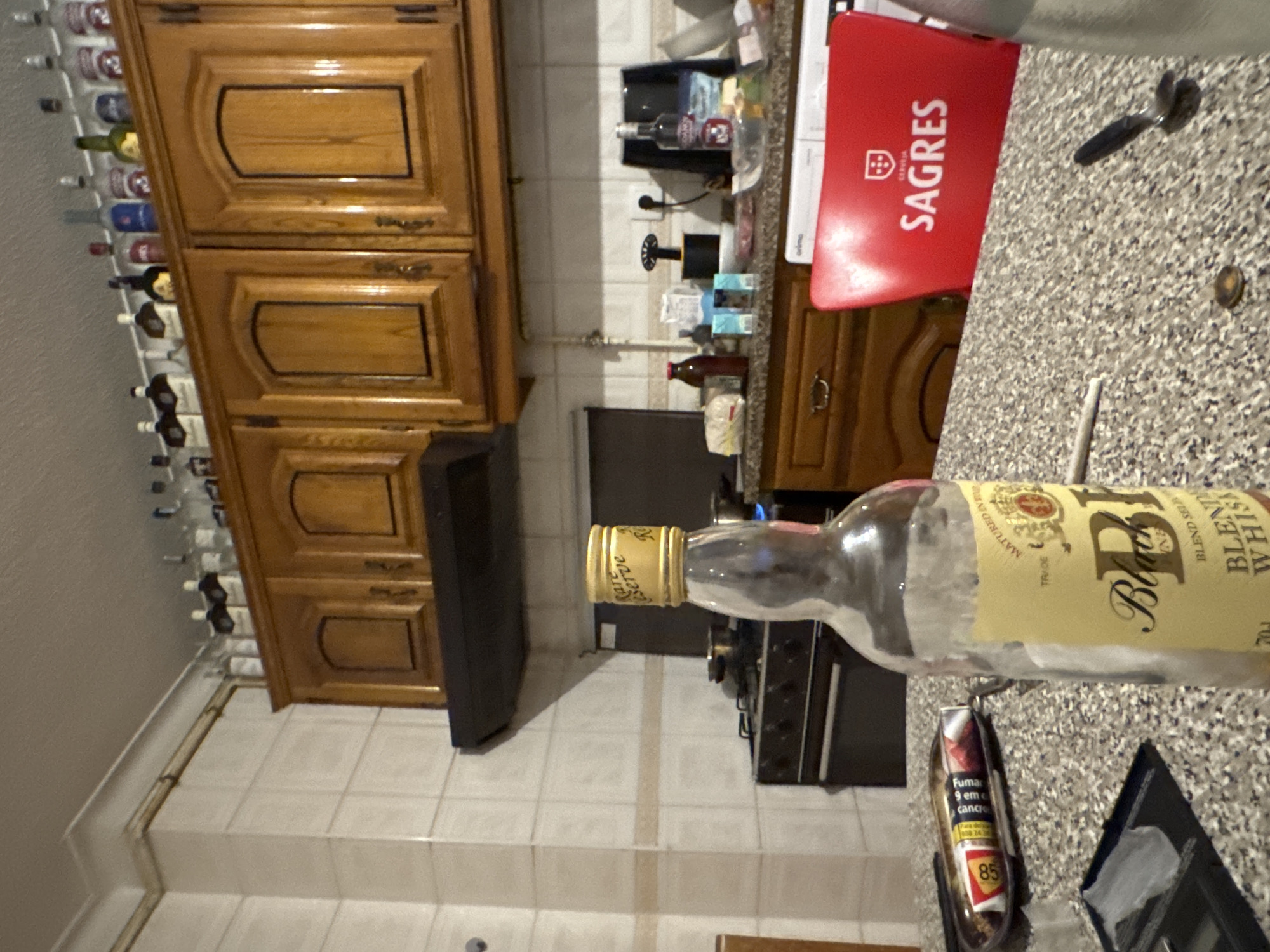 This image depicts the interior of a domestic kitchen in Bragança, Portugal. The primary focal point is a table in the foreground, featuring an almost empty bottle of "Black" blended whisky with a "Rare Reserve" gold cap. Adjacent to the whisky bottle are a rolled cigarette (joint) and a dark, circular object resembling a coin or bottle cap. A silver spoon with a black handle rests nearby. Also on the table is a black rectangular container with white residue, and a pack of cigarettes displaying a Portuguese health warning.
In the midground, a cluttered kitchen counter is visible, containing various items including bottles (one red-capped, one silver-labeled), bags, and boxes. A red object, identified as a cooler or chair back, displays the "SAGRES" beer logo and "CERVEJA" (Portuguese for beer). To the left, a dark-colored stove with four burners is present, with one burner on the right exhibiting a blue flame and pots resting on top. Above the stove is a dark range hood. The walls are tiled in white.
The background features numerous wooden upper cabinets with arched decorative panels. On top of these cabinets, a collection of approximately 20-25 empty or partially full liquor bottles of various shapes and colors are displayed. The ceiling is cream-colored. No individuals are visible in the scene.