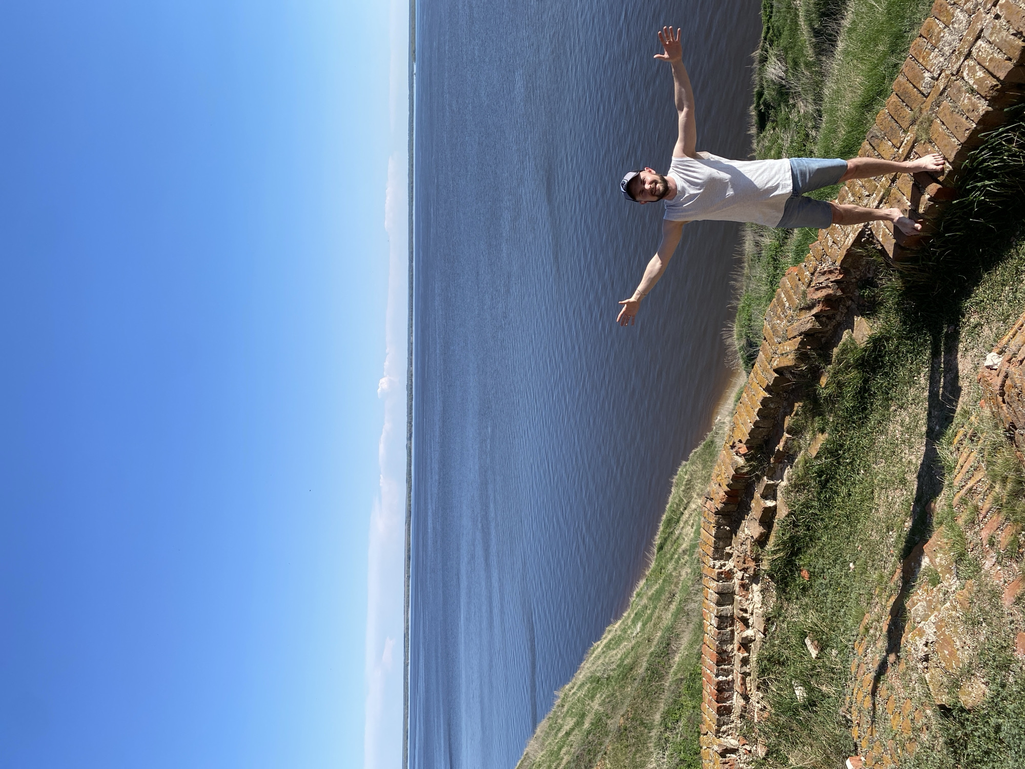 A low-angle shot depicts a light-skinned person standing barefoot on a weathered brick structure adjacent to water. The person wears a light-colored t-shirt, dark shorts, and a hat, and holds their arms outstretched. The water body fills the middle portion of the image, with the clear blue sky occupying the left side. The brick structure is topped with grass and vegetation. The photograph was taken in Шуран, Russia.