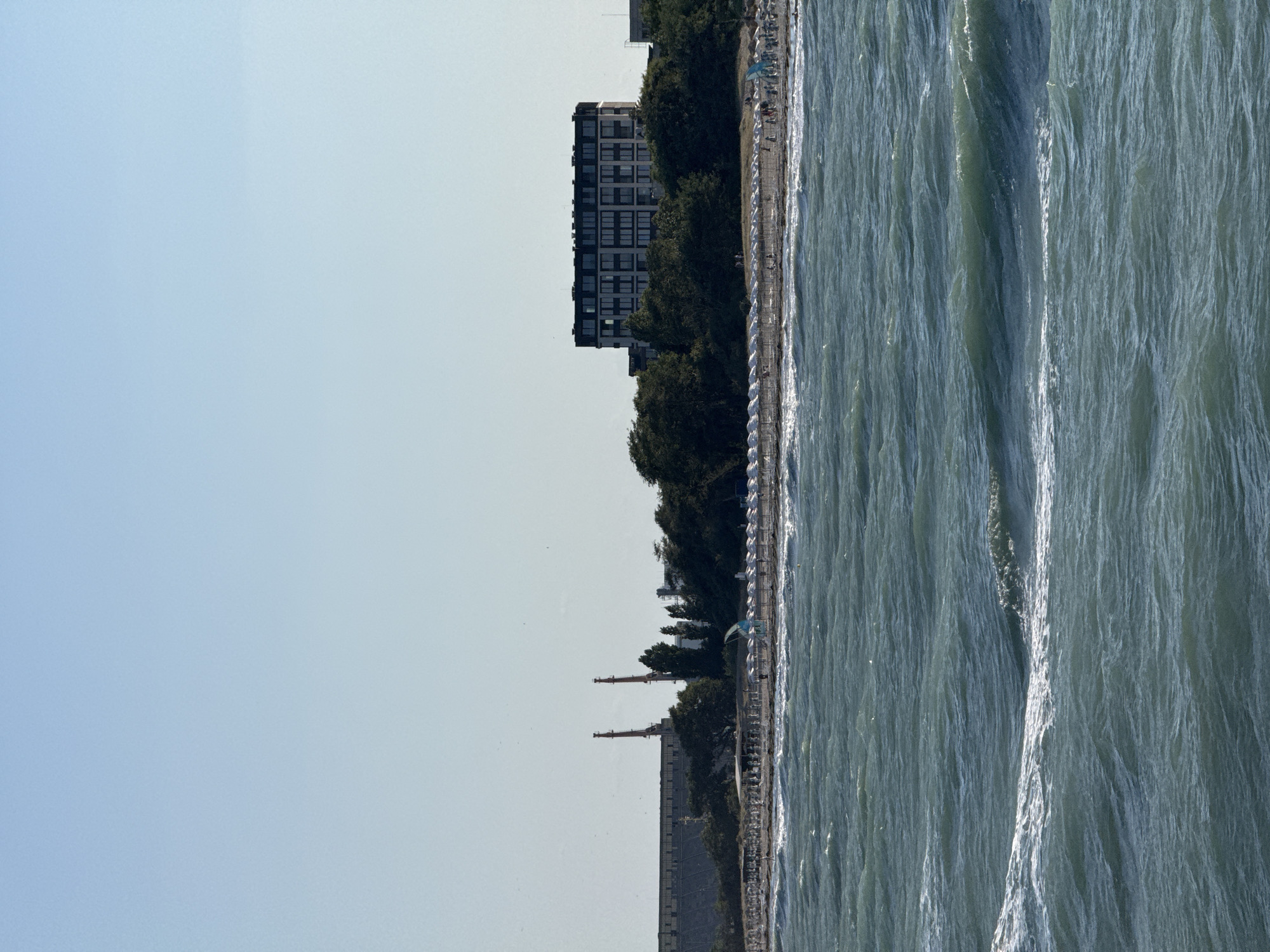The image depicts a scenic view of the Black Sea coast in Burgas, Bulgaria. The foreground is dominated by choppy, dark teal water with whitecaps. In the middle ground, a sandy beach is lined with rows of white umbrellas and lounge chairs, suggesting a popular recreational area. Several small figures of people are visible on the beach, some walking and others possibly sunbathing. A light blue and white kite is set up on the sand. Behind the beach, dense green trees form a backdrop for a modern, multi-story building with a dark facade and many windows. Further in the distance, a large industrial structure with two tall, red and orange chimneys is visible on the left side of the frame. The sky is clear and a pale blue, indicating a bright day. The overall scene captures a coastal landscape with a blend of natural beauty and urban development.