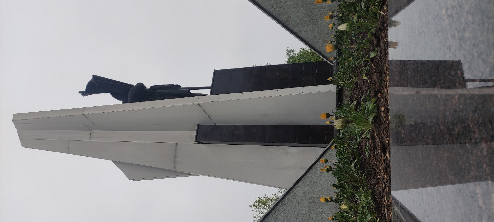 The image depicts a monument in Fryazino, Russia, featuring a dark sculpture of figures holding a flag atop a white, angled structure. Adjacent to the monument are flowerbeds with yellow flowers. A gray reflective surface, likely part of the memorial, reflects the sky. The day appears overcast.