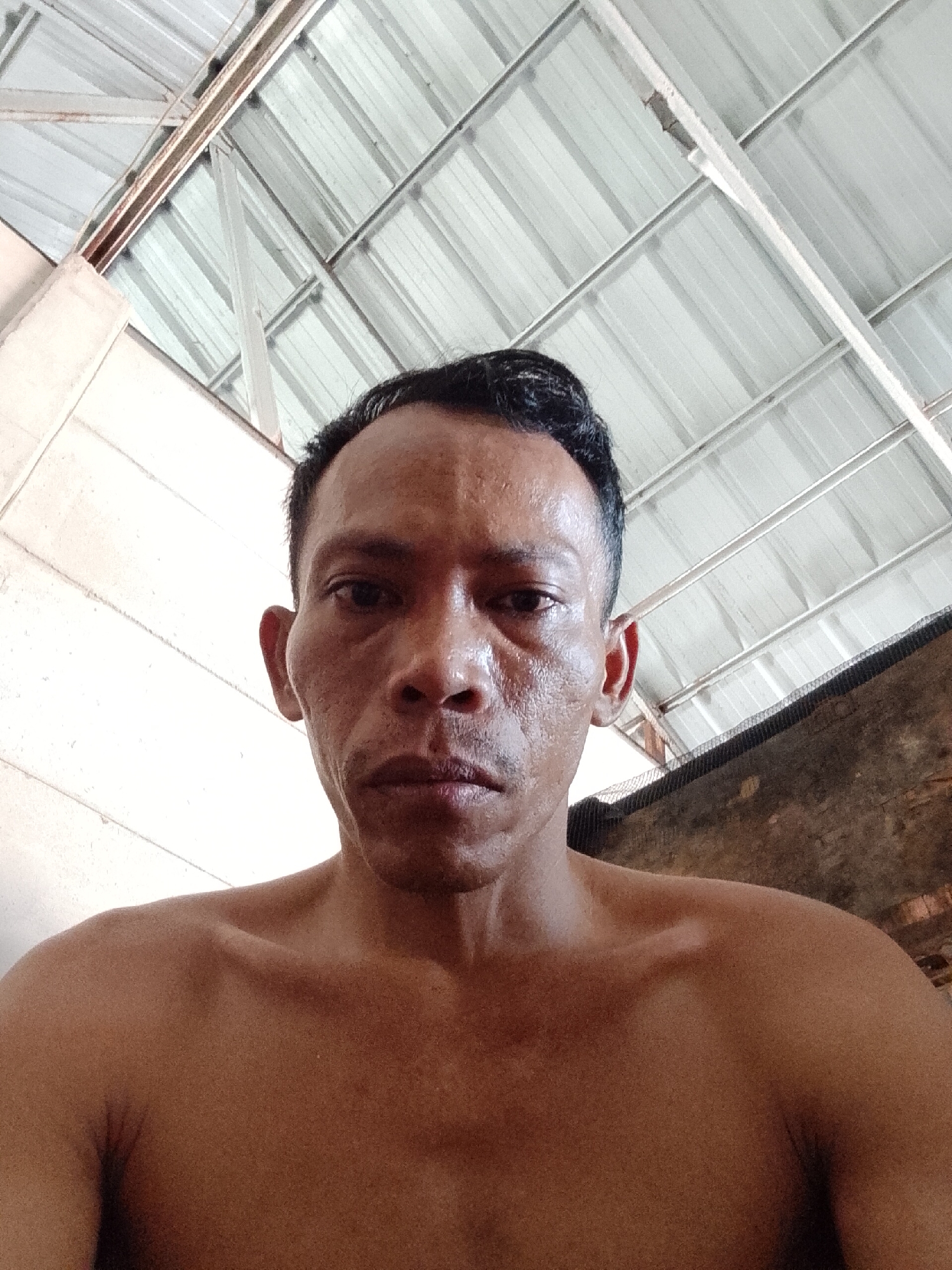 A medium close-up shot taken from a low angle features a shirtless middle-aged man. He has dark hair with a receding hairline. His skin tone is brown, and his face exhibits signs of aging, including wrinkles around the eyes and mouth. He has a serious expression.
The background consists of a white corrugated metal roof with visible support beams, indicating an indoor or sheltered space. A wall painted white is visible on the left side of the frame. On the right side, there appears to be a brick or concrete wall with a dark netting or mesh material attached to it.
The photograph was likely taken in Bukit Ketok, Indonesia, based on the prompt's specification. The environment suggests a residential or semi-industrial area.