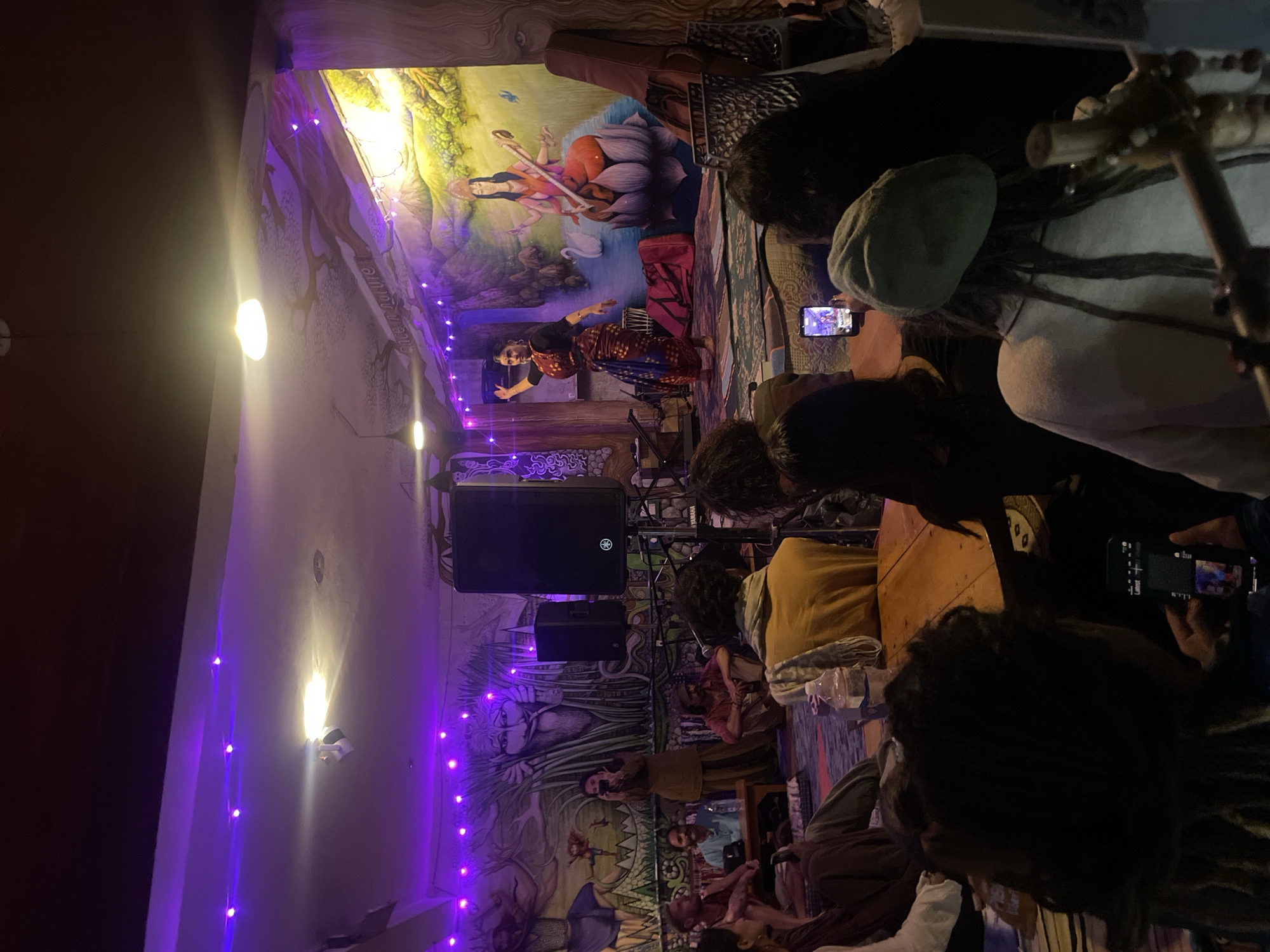 The image depicts an indoor event with a stage set up at one end. A woman in traditional Indian attire is performing a dance on the stage, characterized by a colorful sari and arm movements. The backdrop of the stage features a mural with a depiction of a deity seated on a lotus flower. On the stage, there are also musical instruments and bags. In the foreground and to the sides, an audience is seated on the floor and on low seating, watching the performance. Many audience members are looking towards the stage, and some are holding up their phones to record or take pictures. The room is decorated with purple string lights and murals on the walls that appear to have a spiritual or artistic theme. A large speaker is positioned near the stage. The lighting is dim, with purple and white lights illuminating the space. The overall atmosphere suggests a cultural performance or gathering in a community space or small venue. The location is not specified as Haini, India.