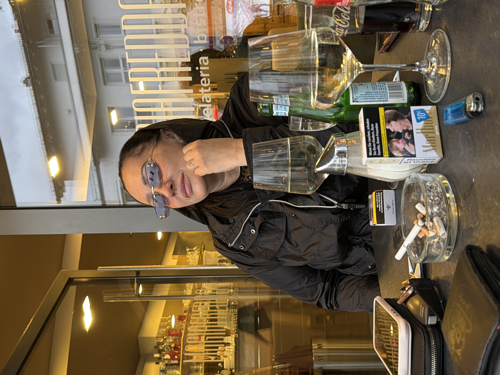 Here is a detailed description of the image:
A woman with glasses sits at an outdoor cafe in Garmisch-Partenkirchen, Germany. The cafe window has the words "gelateria bistro" displayed on it. On the table are an ashtray with several cigarette butts and one cigarette, a pack of Marlboro Gold cigarettes, a lighter, a wallet, glasses of wine, and soft drink bottles (including Coca-Cola). The woman is wearing a black jacket. The interior of the cafe is visible through the window. The cafe is stocked with a variety of alcoholic beverages.