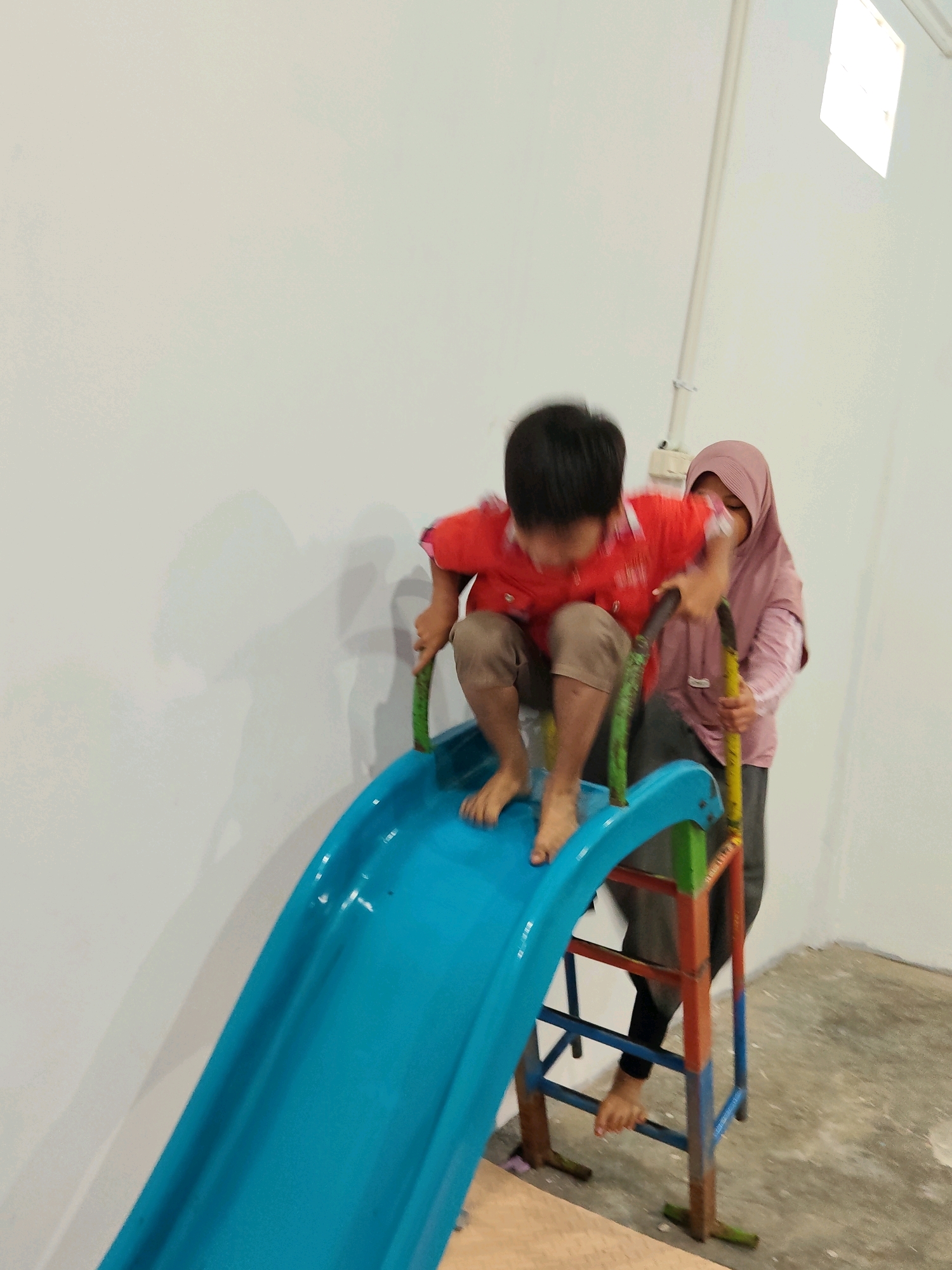 An image taken in Purwakarta, Indonesia, shows a young boy in a red shirt and tan shorts preparing to slide down a bright blue slide. He is holding onto the green supports of the slide with his hands. Behind him, a woman wearing a pink hijab and a pink long-sleeved shirt stands near the top of the slide, appearing to assist or supervise. The slide is part of a multi-colored metal structure with red, blue, and green components. The scene is indoors, with a white wall in the background and a concrete floor. A light fixture is visible on the upper right portion of the wall.