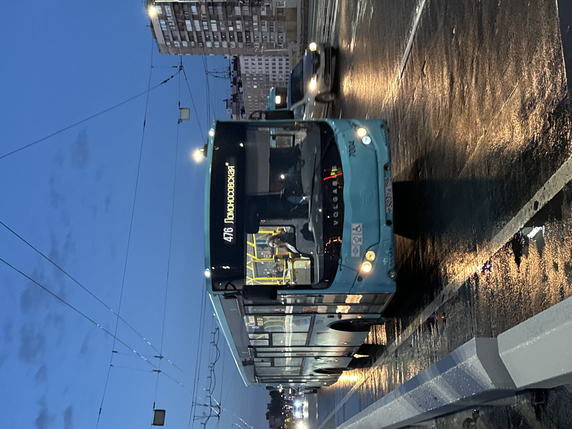 The image depicts a light blue public transportation bus, identified by the number 7004 and the route number 476 "Lomonosovskaya", moving on a wet street in Saint Petersburg, Russia. The bus is the primary subject, facing forward and slightly to the right, with its headlights illuminated. Inside the bus, a female passenger with dark curly hair, wearing a dark jacket, is visible in the front passenger area. Another passenger, a child, is also discernible behind her.
The bus is operating on a multi-lane road, evident from the lane markings and the presence of another silver car to the right, also with its headlights on. The road surface is wet and reflects the streetlights and vehicle lights, suggesting it has recently rained or is raining. Overhead, electric wires for the bus are visible against a twilight sky, which is a deep blue with some faint clouds.
In the background, on the right side of the image, a tall, multi-story residential building is present, characteristic of urban architecture. The overall environment is an urban street scene at dusk or dawn, with the low light conditions contributing to the reflections on the wet road. The scene is captured from a low angle, slightly below the bus's midline.