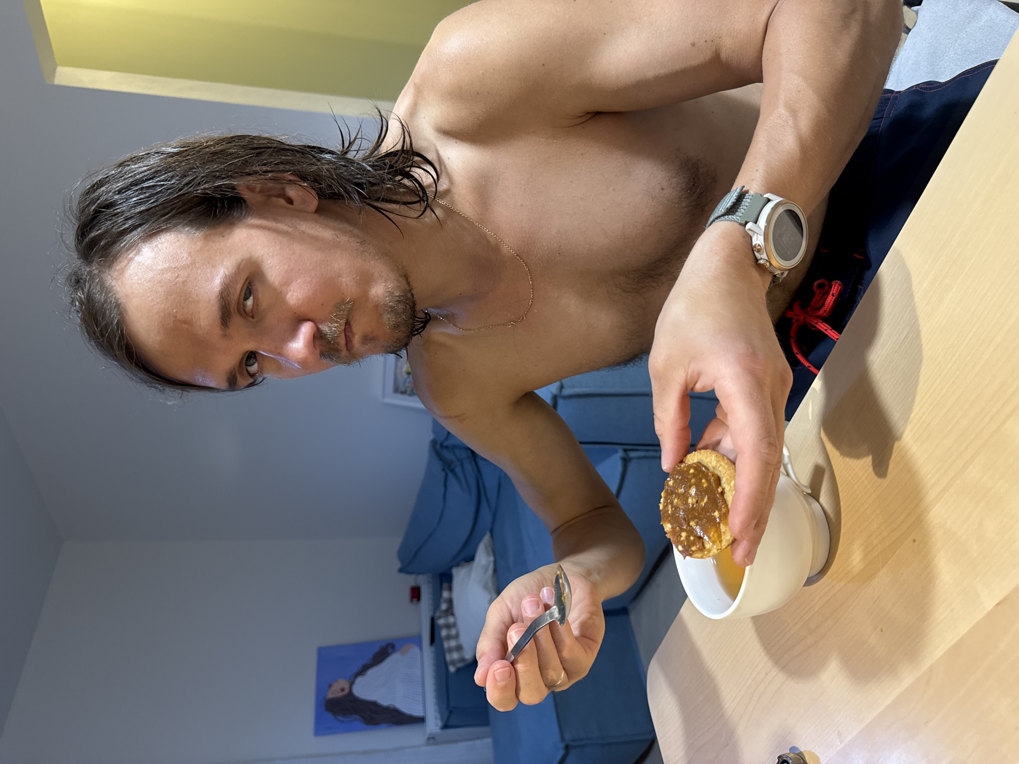 An indoor, waist-up shot, rotated 90 degrees clockwise, captures a shirtless man in what appears to be a home in Izhevsk, Russia. He has medium-length, dark hair, a mustache, and a short beard, and looks directly at the camera with a neutral expression. A thin gold chain hangs around his neck, and a prominent white and gold smartwatch with a grey strap is on his left wrist.
In his right hand, the man holds a round biscuit or cookie topped with a thick, textured, brownish spread, possibly peanut butter or a caramel-nut mixture. He positions it above a white ceramic mug containing a light-colored liquid, suggesting he is dipping the biscuit or about to consume it. His left hand holds a small spoon or utensil.
The background features light-colored walls, a large blue sectional sofa, and a framed painting on the wall depicting a stylized figure in white against a blue background. A light wooden tabletop occupies the foreground and side of the frame. The lighting is artificial. No visible text is present in the image.