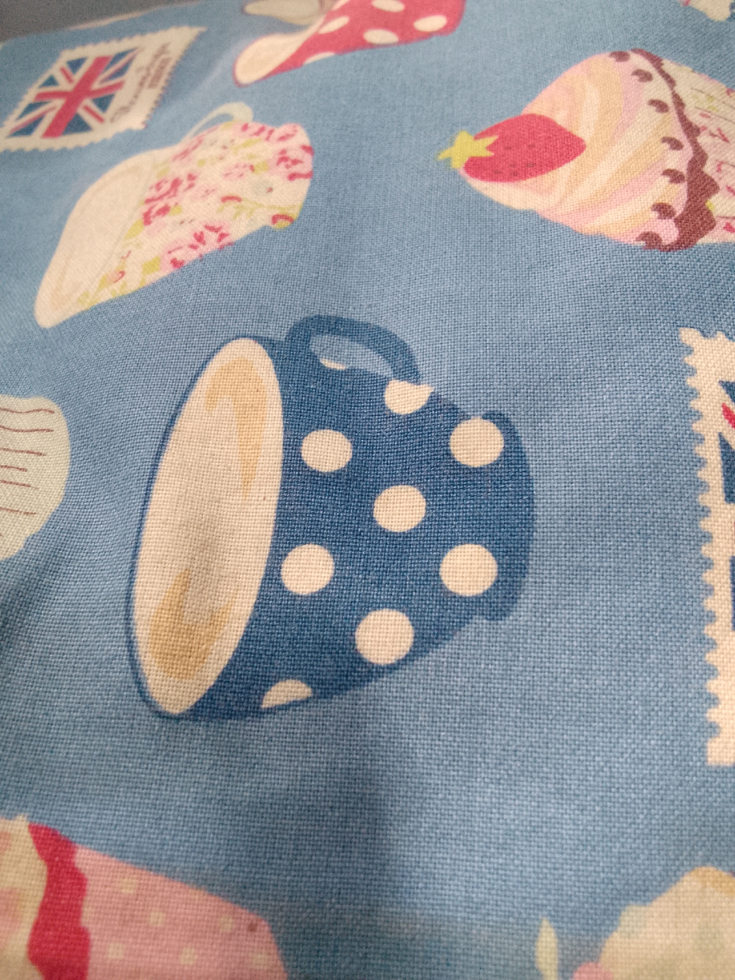 The image displays a close-up, slightly angled view of a blue fabric with a pattern of teacups and stamps. The fabric has a visible woven texture. The teacups are depicted in a playful, cartoonish style, with one prominent blue teacup adorned with white polka dots, shown from a side profile. Inside this teacup, a light brown substance, possibly tea or coffee, is visible. Other teacups in the background feature floral patterns in pink and white. The stamps, like the Union Jack, are also part of the repeating design. The lighting is soft, casting a slight shadow on the teacup. There is no indication of the city of Karangan, Indonesia, in this image, as it is a close-up of fabric and not an outdoor or environmental scene.
