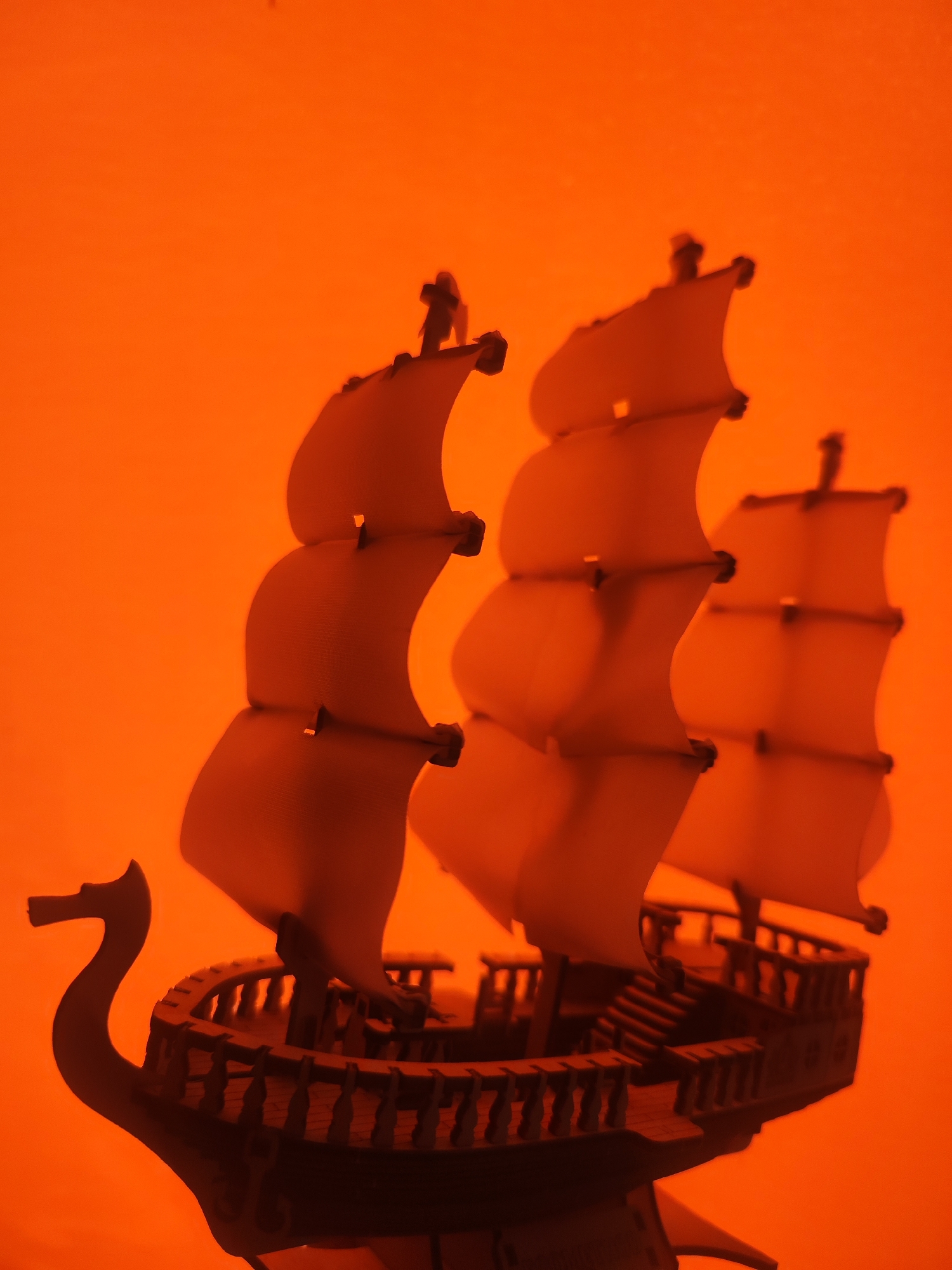 The image depicts a model ship, likely a galleon or similar sailing vessel, against a solid orange background. The ship has three masts, each with multiple sails. The hull of the ship is detailed with a railing and other architectural elements. The image appears to be a studio shot with controlled lighting. There is no indication of the image being taken in or near the city of Chaikovsky, Russia. The image includes only the model ship and the background, so there are no people or interactions to describe. The overall environment is artificial, suggesting the model is indoors.