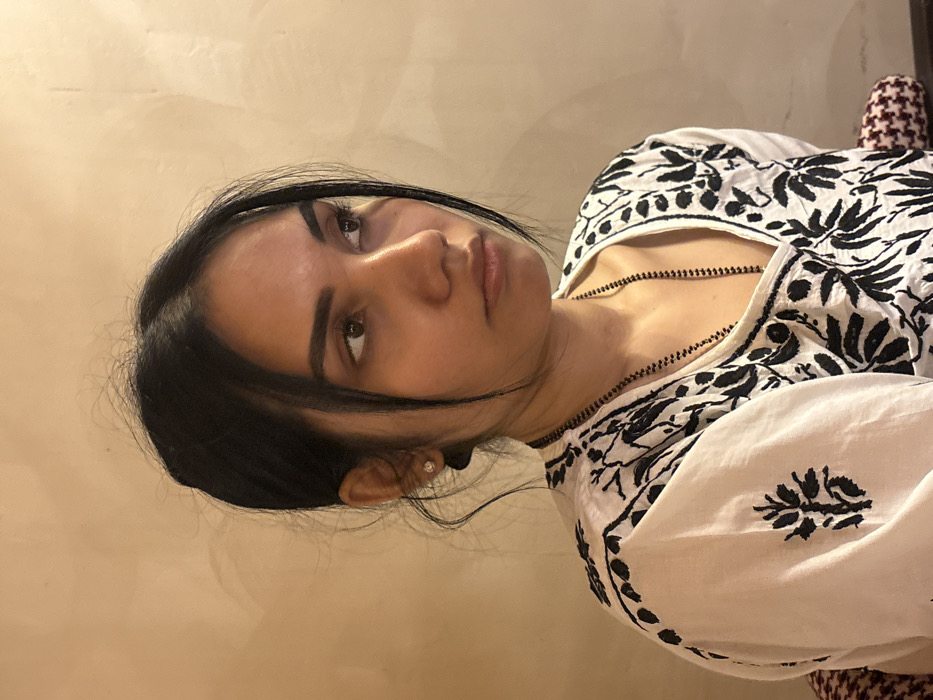 A young adult female, appearing to be of South Asian descent, is depicted in a three-quarter profile, looking towards the right side of the frame. She has dark hair, styled in a bun with loose strands framing her face, dark brown eyes, and a neutral expression. She is wearing a white garment with extensive black embroidered floral/leaf patterns, featuring a V-neckline. A thin, long black beaded necklace hangs around her neck, and a small, circular, light-colored stud earring is visible in her left earlobe. The background consists of a plain, light beige textured wall. A small portion of a red and white patterned fabric, possibly a cushion, is visible in the lower right corner. The scene takes place in an indoor setting within the city of Howrah, India.
