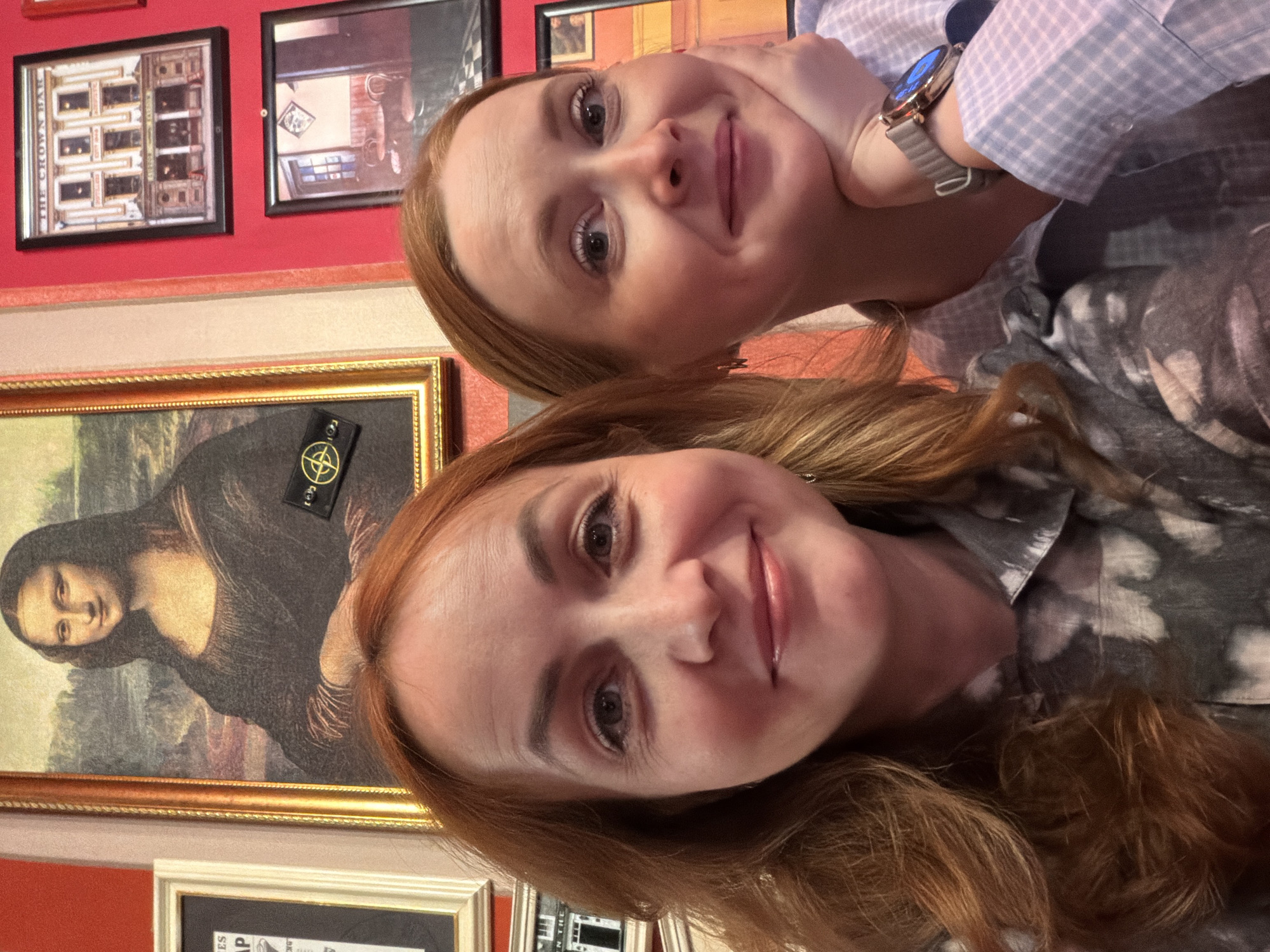 Two women are posing for a selfie in front of a wall decorated with framed pictures, including a rendition of the Mona Lisa with a Stone Island patch. The women have red hair and are smiling. One woman is wearing a light blue checkered shirt and a watch. The background is a red wall with framed pictures. The location is Chaikovsky, Russia.