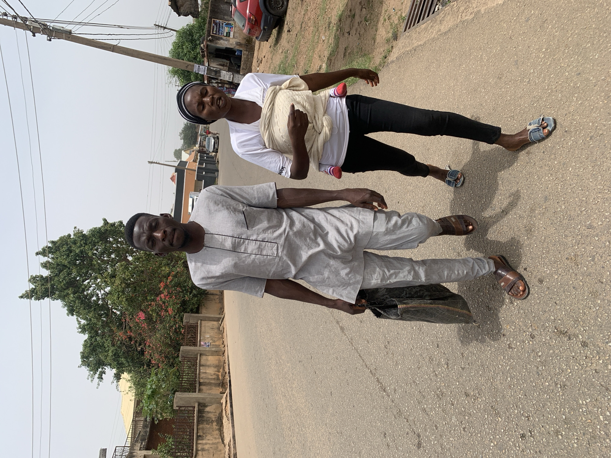 A medium shot captures a man and a woman walking on a paved road. The woman is carrying a baby secured in a wrap, and she is wearing a white t-shirt, black leggings, a black headband, and sandals. The man is dressed in a light grey outfit and brown sandals, and he is carrying a black bag. They appear to be in a residential area, with buildings, a tree with flowers, and utility poles visible in the background. The location is in the city of Jos, Nigeria.