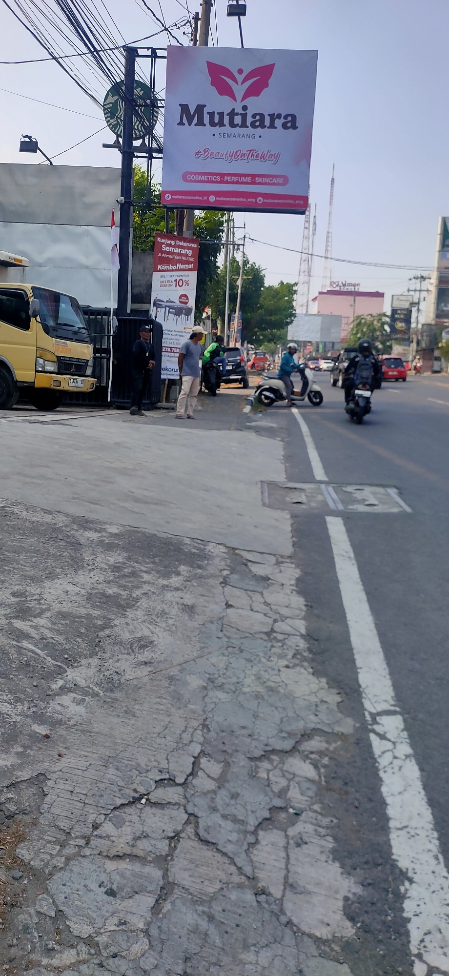 This is a street-level, eye-level, medium shot photograph taken outdoors in Semarang, Indonesia. The image captures a busy urban scene, likely on a commercial street.
In the foreground, a cracked and weathered concrete sidewalk runs diagonally across the frame from the bottom left corner upwards towards the right. The sidewalk is textured with various shades of grey and shows signs of wear and tear. A white line marking a traffic lane is visible on the right side of the frame, indicating the edge of the road.
Further back, a yellow truck is parked on the left side of the image, partially obscuring a black metal gate. Two individuals are standing near the gate: one person in dark clothing, appearing to be a security guard, stands alert, while another person in a light-colored shirt and pants stands facing the street.
Across the street, several vehicles are present, including motorcycles and cars. One motorcycle has fallen on its side in the middle of the road, with a rider nearby. Other motorcycles and cars are in motion or parked. In the background, a large billboard for "Mutiara" advertising cosmetics, perfume, and skincare is prominently displayed on a pole. The billboard features the brand's logo and the hashtag "#BeautyOutTheWay".
Behind the billboard and other structures, tall telecommunication towers are visible against a clear blue sky. The overall environment suggests a lively commercial area with ongoing traffic and pedestrian activity.