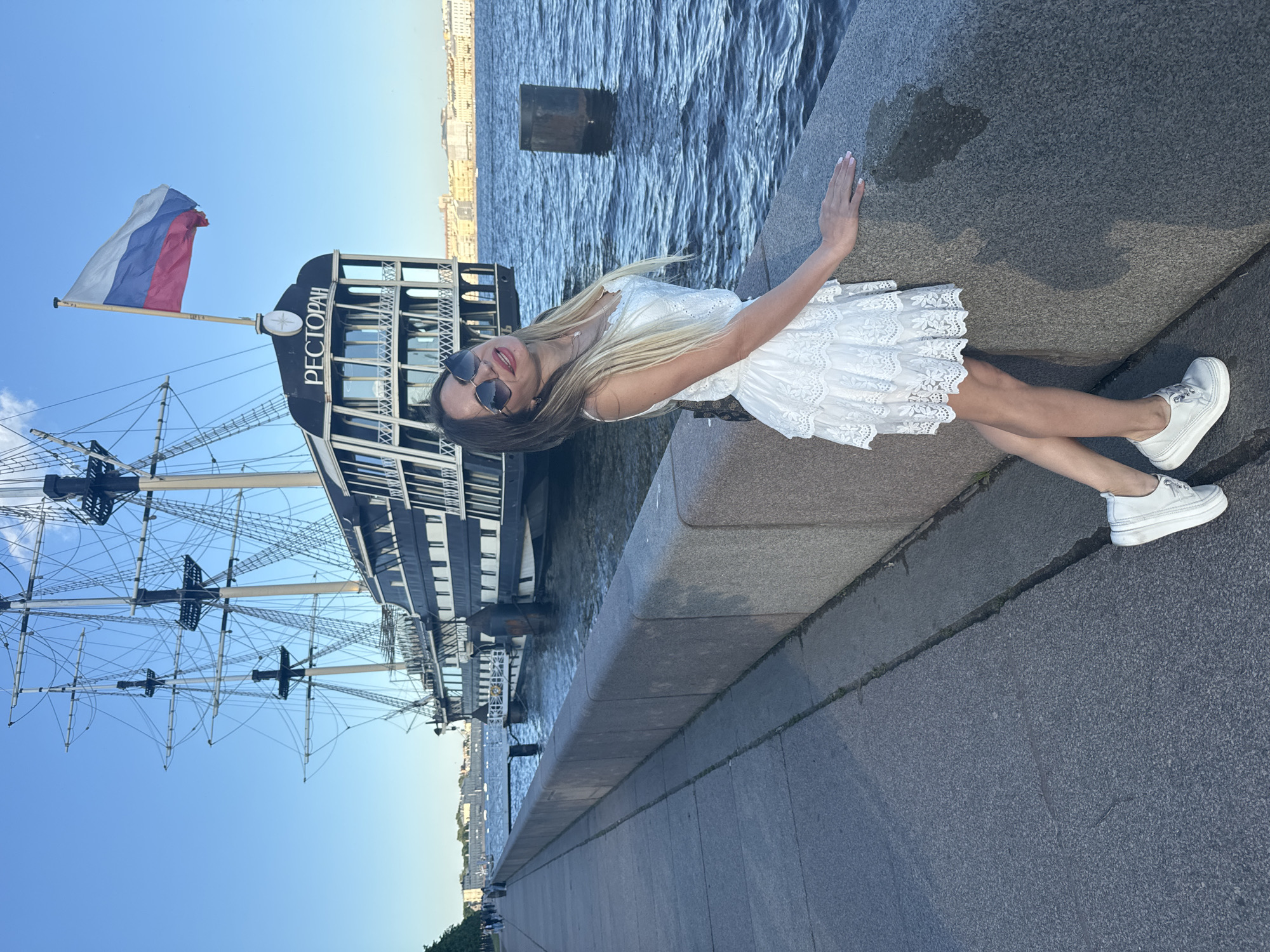 A young woman in a white dress and white sneakers is posing by a stone embankment in Saint Petersburg, Russia. She is leaning on the embankment and looking at the camera with a smile. Behind her, a large ship with tall masts and a Russian flag is docked on the Neva River. The ship is labeled with the word "РЕСТОРАН" (Restaurant). In the background, the cityscape of Saint Petersburg is visible. The sky is clear and blue, and the water is calm. The image appears to have been taken on a sunny day.