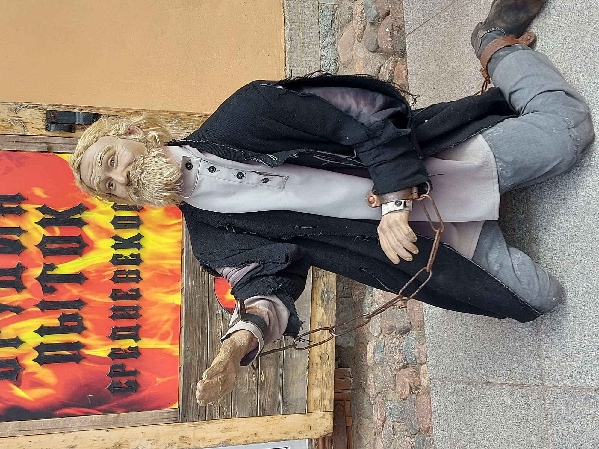 A full-length image depicts a chained mannequin of a man. He has a light-colored beard and hair and is wearing a long-sleeved grey button-up shirt under a black jacket with ragged edges. His lower body is grey and he's wearing a boot. Chains are attached to the man's wrists and ankles. The mannequin is adjacent to a sign with orange and black text written in Cyrillic. The background shows a stone wall and a beige wall. The mannequin is located in Saint Petersburg, Russia.