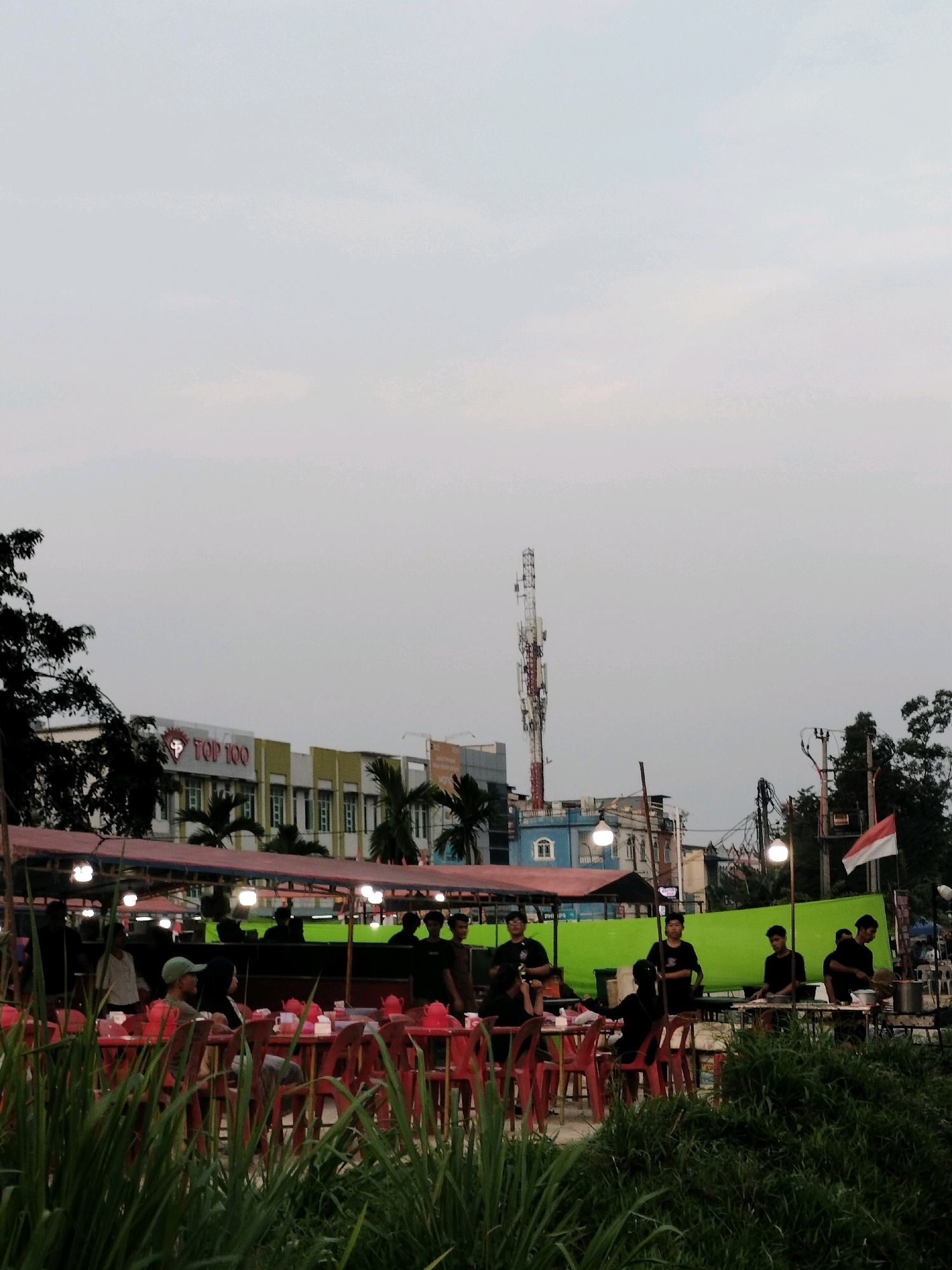 This is a photograph of an outdoor dining area, likely a food stall or market, in Batam, Indonesia. In the foreground, out-of-focus green grass is visible, suggesting a natural setting adjacent to the dining area. Behind the grass, rows of red plastic tables and chairs are set up, some occupied by people. Several individuals are seated at the tables, engaged in conversation or dining. In the middle ground, under a canopy with lights, more tables are visible, with people standing and working, possibly preparing food. A bright green tarp serves as a backdrop for some of these stalls.
In the background, a row of buildings is visible. One building features a prominent red and white sign with "TOP 100" emblazoned on it, indicating a business or establishment. Further in the background, a tall communication tower with several antennas stands prominently against the pale, overcast sky. Other structures, including what appears to be a blue building with white window frames and power lines, are also present. An Indonesian flag is flying from a pole on the right side of the image. The overall impression is of a lively, informal dining scene in an urban environment.