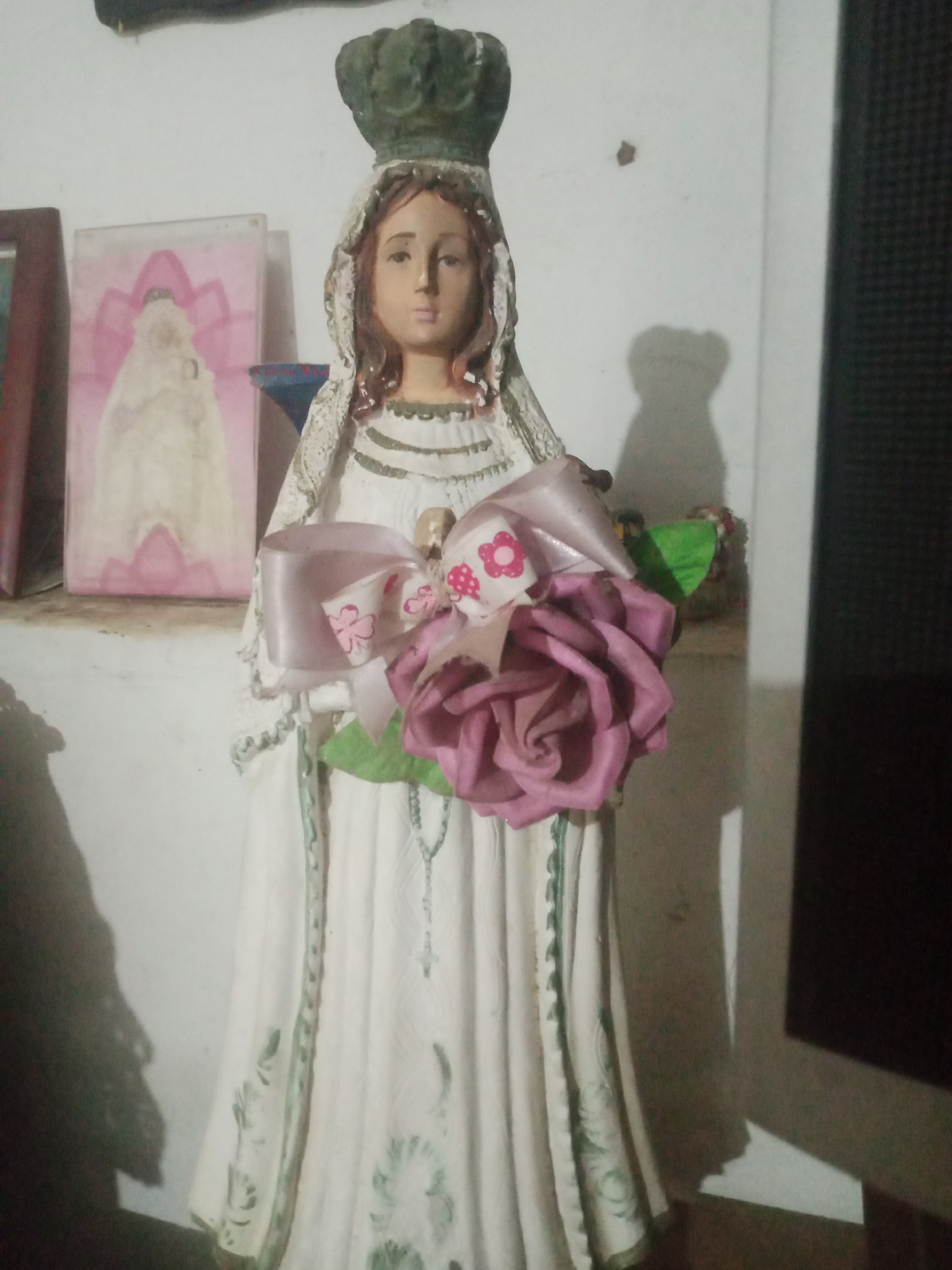 A statue of the Virgin Mary stands on a shelf against a white wall. The statue is dressed in a white gown with green trim and holds a bouquet of artificial flowers, including a large pink rose, and a rosary drapes from its hand. To the left of the statue, a framed painting depicts a figure in a white gown and pink headdress. The statue and painting appear to be placed on a shelf in a home or religious space. The lighting is dim, casting a shadow of the statue on the wall. No individuals are present in the image. The background consists of a white wall and a dark object to the right. The context of Santa Ana, Venezuela is not discernible from the image itself.