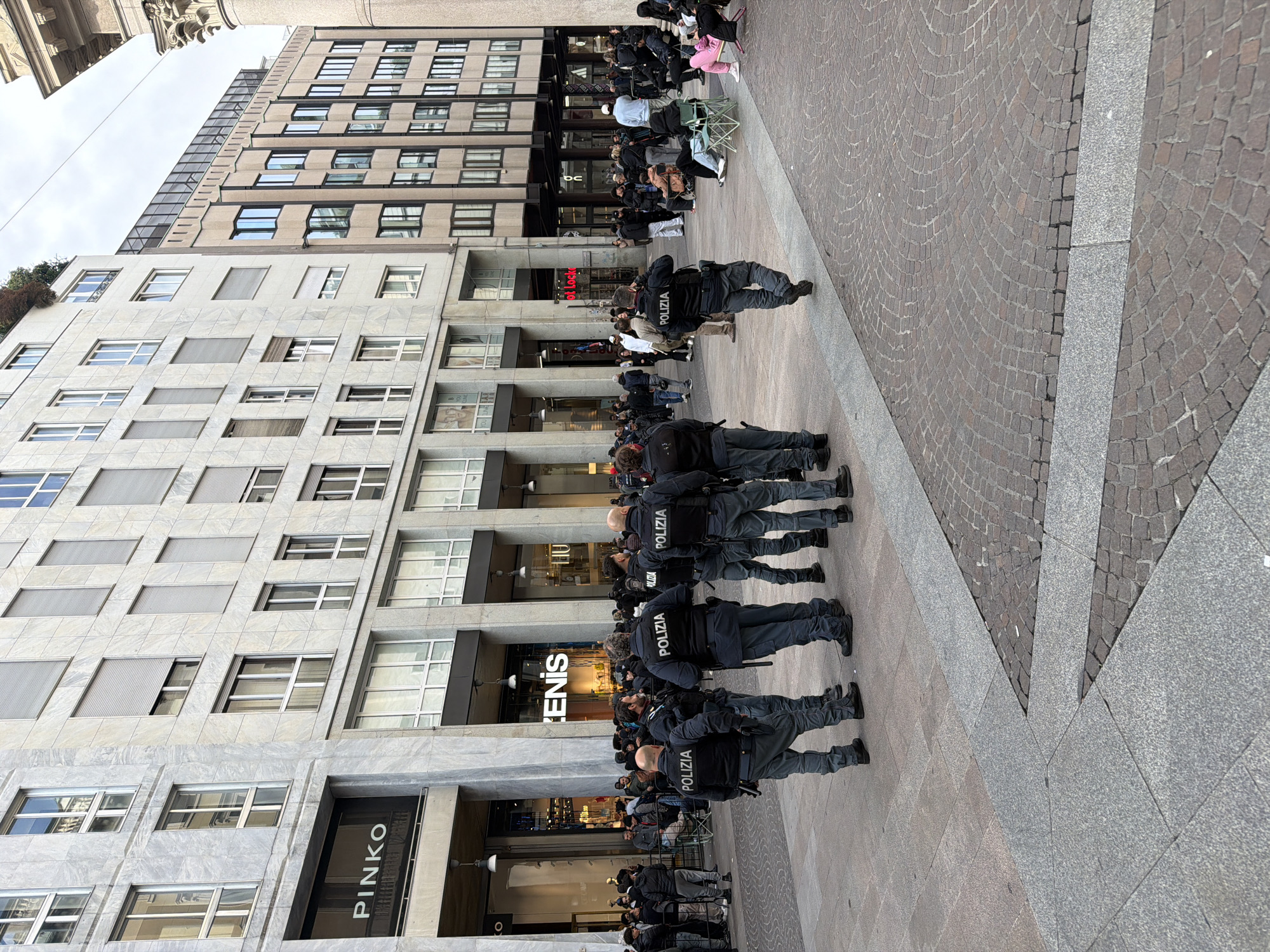 The image depicts a scene in Milan, Italy, with a line of Italian police officers ("Polizia" is visible on their backs) standing shoulder-to-shoulder facing a crowd of people seated and standing. The officers are wearing dark uniforms. The crowd is a mix of individuals of various ages and appearances. Behind the crowd and the police officers are buildings with storefronts, including "PINKO" and "DENIS". The ground is paved with patterned stone tiles. The sky is overcast.