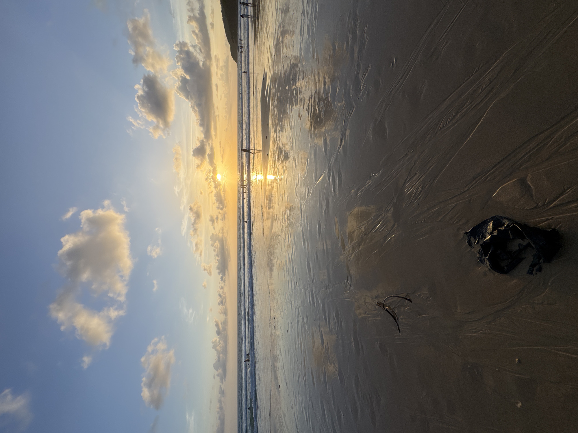 This image captures a sunset on Karon Beach in Karon, Thailand. The sky is a blend of blues and oranges, with scattered clouds catching the golden light of the setting sun. The sun itself is visible on the horizon, casting a bright reflection on the wet sand and the gentle waves. Several people are present on the beach, some wading in the water and others walking along the shore. The foreground of the image focuses on the wet sand, marked with ripples and textures from the receding tide. Two dark objects, likely debris, are visible on the sand in the foreground. The overall scene depicts a serene beach atmosphere at dusk.