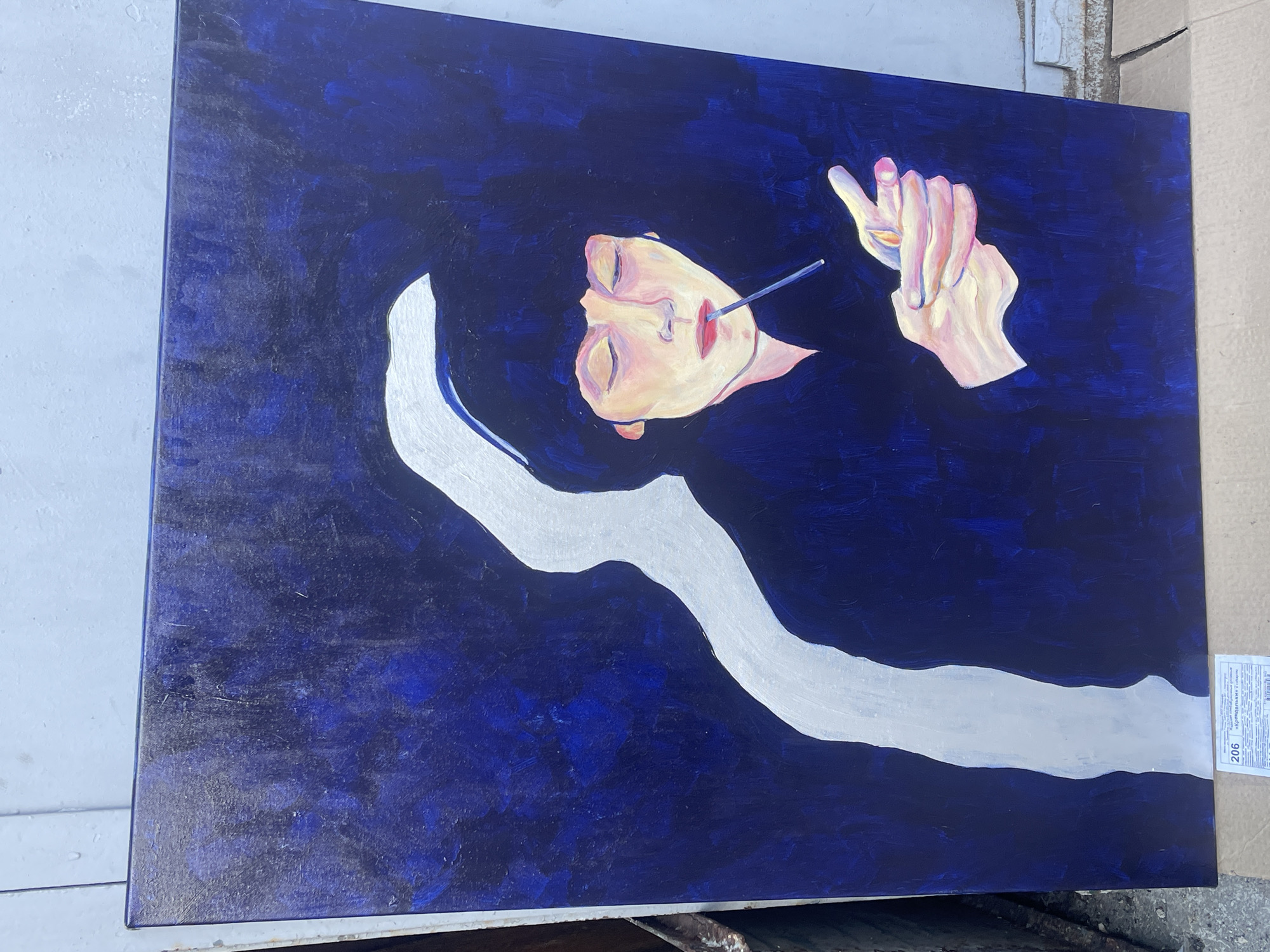 A painting depicts a person with pale skin and red lips, lying in dark blue water. A silver form resembles a lock of hair or a stream. A cigarette is held between the person's lips and fingers. The surrounding environment includes parts of a building in Yerevan, Armenia, and possibly a cardboard box, which are outside the main focus of the artwork.