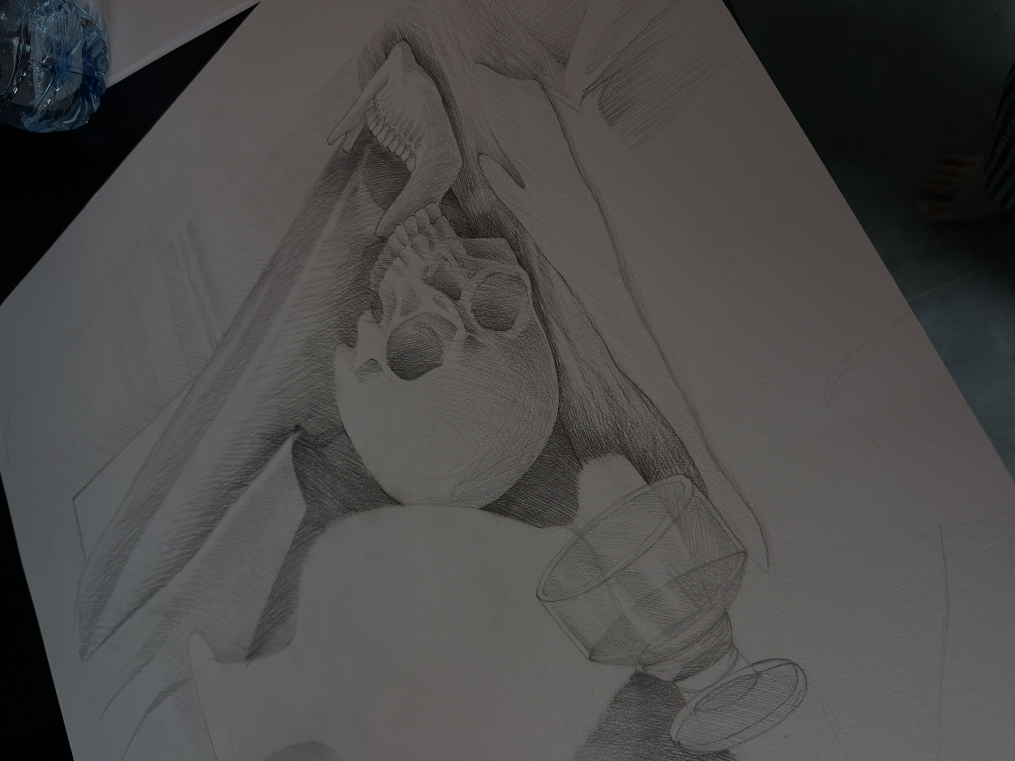 This is a black and white sketch on paper, showing a still life composition featuring a skull, a jawbone, a draped cloth, and a glass. The skull is centrally located and is in an upside down orientation, with the jawbone set above it. The cloth is draped around the skull, creating shadows and highlights. The glass is located in the lower right corner of the image, slightly angled. A water bottle is present in the upper left corner outside of the main composition. A foot can be seen in the upper right corner of the frame. The artwork has been executed with shading and attention to detail. The location is unknown.