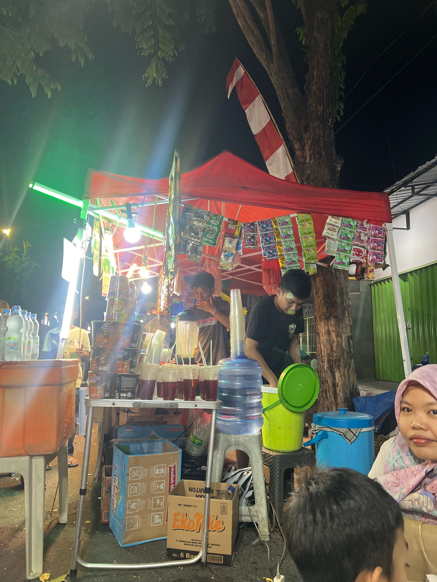 An outdoor night scene in Kecamatan Pati, Indonesia, depicts a street food stall under a red canopy. Several people are present, including two young men operating the stall and a woman in the foreground. The stall is laden with various products, including bottles of water, stacks of instant noodle cups, and numerous small packets of snacks hanging from the canopy. Several plastic cups filled with a dark liquid are arranged on a table, likely beverages being served. Behind the stall, a large tree with exposed roots is visible, and a red and white Indonesian flag is attached to its trunk. The ground appears to be paved. The lighting is a mix of artificial lights illuminating the stall and ambient darkness. A green plastic bucket and blue cooler are also present.