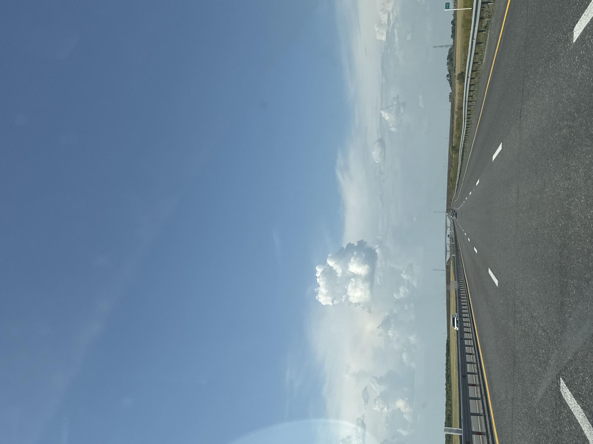 A straight, multi-lane highway stretches into the distance under a vast blue sky dotted with white clouds. The road is paved with asphalt and has clear white lane markings and a yellow line along the right edge. Guardrails line both sides of the highway, separating it from grassy fields on either side. In the distance, a white car is visible in the right lane. Further down the road, a highway sign indicates "142". The environment appears to be rural or undeveloped, with a flat landscape. There are no people clearly visible. The overall scene depicts a travel or journey along an open road on a clear day. The image is captured from a moving vehicle, as suggested by the perspective. The specific location in Алтынды ауылдық округі, Kazakhstan, cannot be definitively determined from the visual information alone, but the landscape suggests a potentially open and vast area common in such regions.