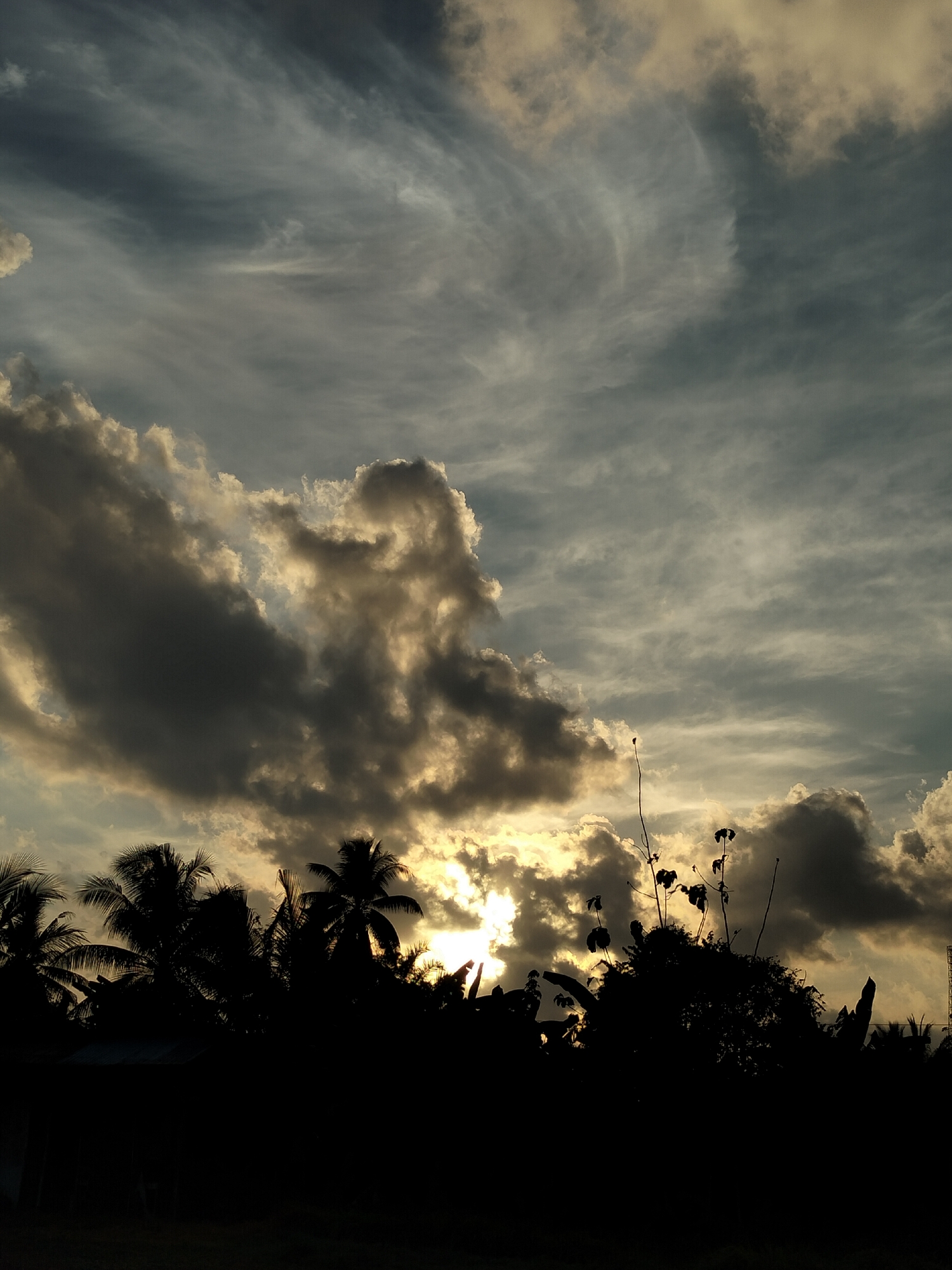 The image depicts a twilight sky over Karang Indah, Indonesia, characterized by dramatic cloud formations illuminated by the setting sun. The foreground is dominated by the dark silhouettes of palm trees and other vegetation, creating a stark contrast against the lighter sky. The clouds are a mix of dark, heavy masses and wispy, lighter streaks, suggesting a dynamic atmosphere. The sun is partially obscured by clouds, casting a warm, golden glow through the breaks. No people or specific structures are clearly discernible in the silhouette, though a hint of a rooftop is visible on the left side of the frame. The overall impression is one of natural beauty and the serene, yet powerful, forces of nature at dusk.