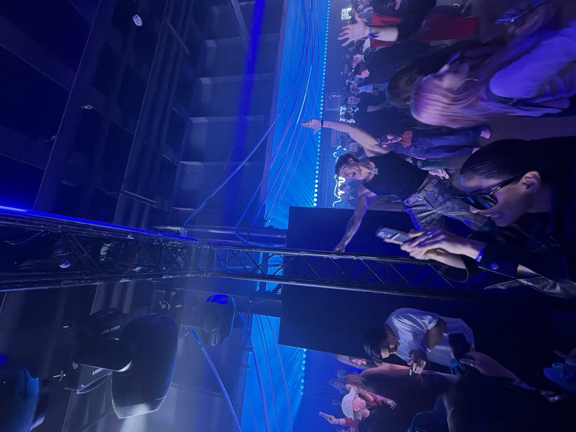 The image shows a crowded event, possibly a concert or club setting, characterized by blue lighting and a stage area. In the foreground, stage lighting equipment is visible. A metal truss structure spans the area. People are present, some holding phones and pointing towards the stage. The crowd appears engaged and lively. The image is rotated clockwise approximately 90 degrees. The location is unconfirmed but has been mentioned to be in Moscow, Russia.