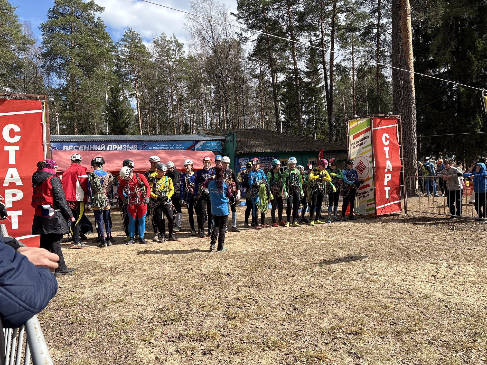 The photograph, taken in daylight, shows a group of approximately 15 individuals preparing for what appears to be an outdoor sporting event in Lesnoye, Russia.
The individuals are wearing helmets and harnesses with ropes, suggesting that the event involves climbing or similar activities. They are standing in a grassy area, some in athletic clothing, and are positioned in front of a banner with the word "СТАРТ" (START), indicating the beginning of the event. The banner also features details in Russian, possibly related to the event's name and organization.
In the background, there are trees, a small building, and a crowd of onlookers behind a barrier. The overall environment suggests a park or wooded area. Some of the participants also appear to be looking at or listening to a woman wearing a blue shirt, who is likely giving instructions or a briefing before the start of the event.
