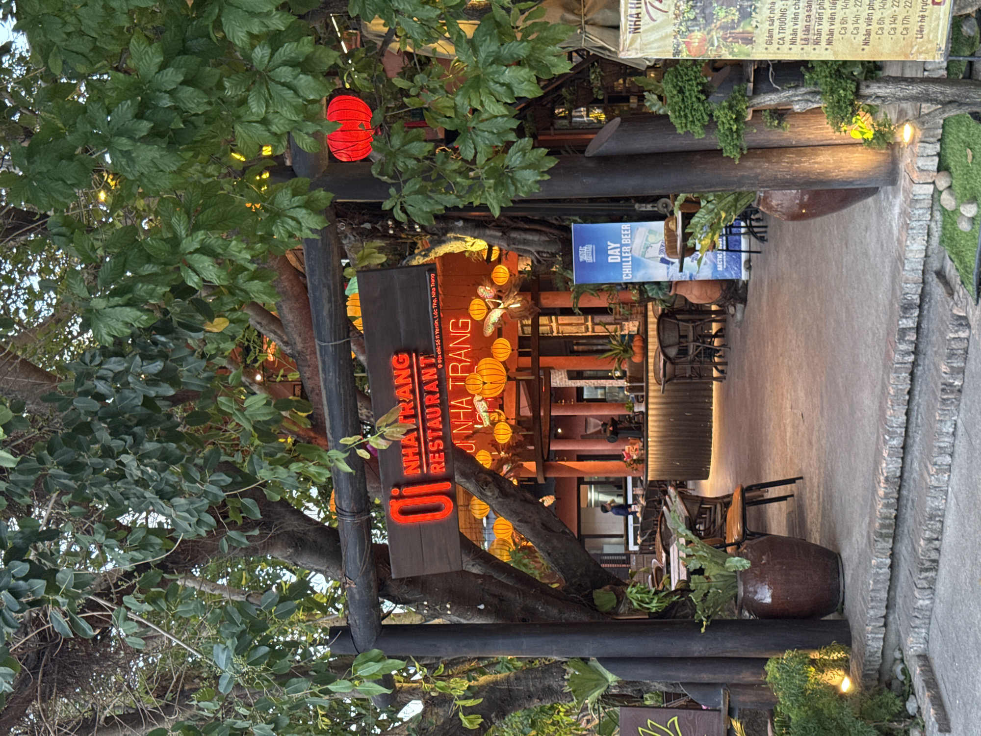 The image shows the entrance to "Oi Nha Trang Restaurant" in Nha Trang, Vietnam. The entrance is framed by thick, dark wooden posts and a horizontal beam, with lush green foliage from large trees hanging overhead and surrounding the area. Red paper lanterns are visible, one hanging prominently on the right side and others glowing warmly behind the wooden sign. The sign itself is dark wood with bright red neon lettering that reads "Oi" and "NHA TRANG RESTAURANT". Below this, smaller text indicates the address: "9 Yersin, Loc Tho, Nha Trang".
The restaurant's interior is visible through the entrance, featuring a warmly lit bar area with a corrugated metal facade and multiple stools. Tables and chairs, made of dark wood and wicker, are set up in the outdoor seating area. A person in a dark blue shirt is visible standing inside the restaurant. To the right of the entrance, a blue poster advertises "DAY CHILLER BEER". Another yellow sign on the far right lists services and prices in Vietnamese. The ground leading to the entrance is paved with concrete and lined with brick steps, with potted plants, including a large ceramic pot with a fern, placed on the steps and surrounding areas. Small rocks and mossy patches are visible on the ground. The overall atmosphere suggests a welcoming and somewhat tropical dining establishment.