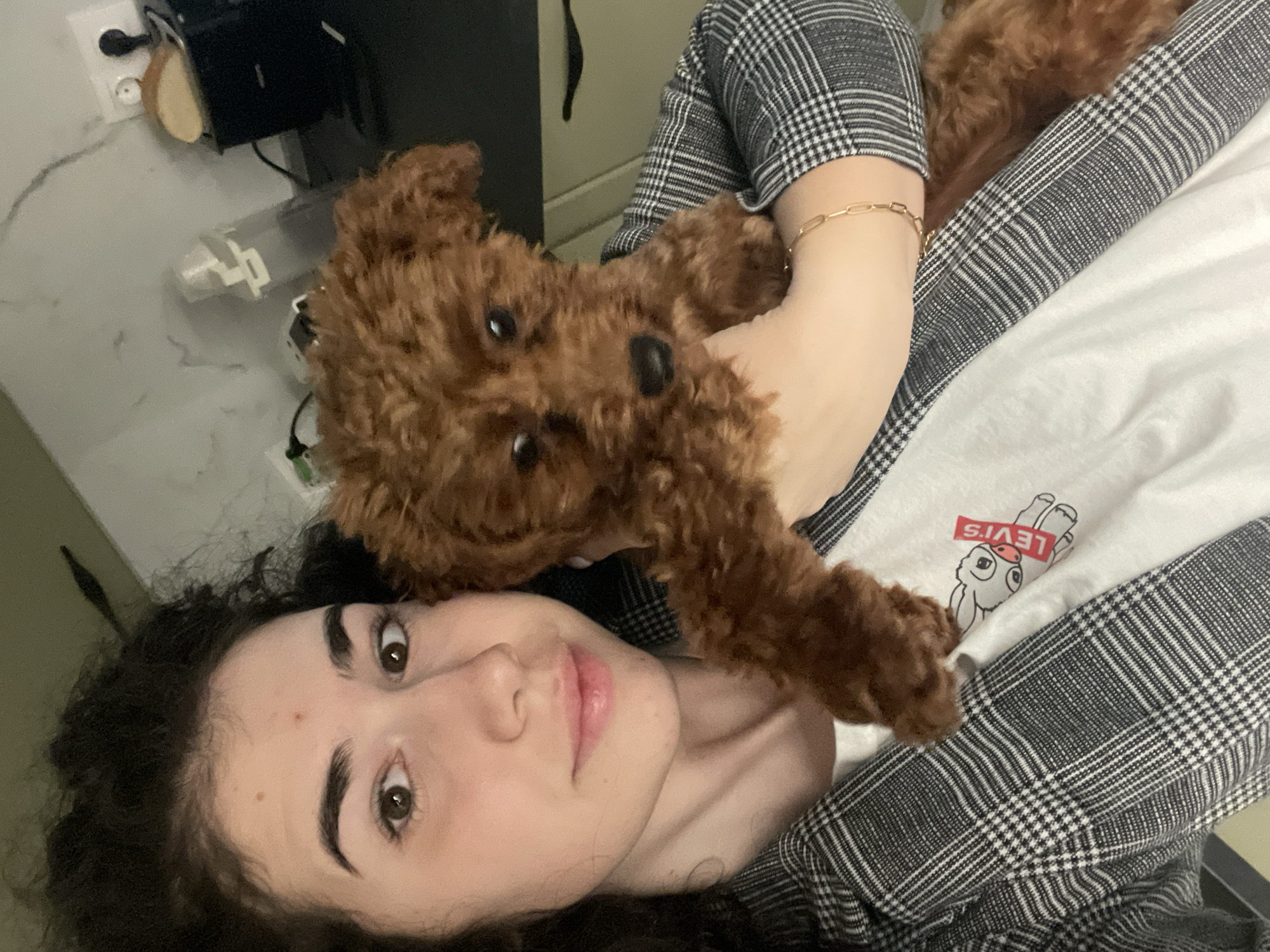A young woman with dark curly hair is holding a brown toy poodle. The woman is wearing a black and white plaid jacket over a white t-shirt with a Levi's logo and a rabbit cartoon. A gold bracelet is visible on her wrist. The background shows a white wall with a power outlet and some electronic devices. This appears to be a domestic setting, likely indoors. It's impossible to pinpoint the exact location in Sofia, Bulgaria without more information.