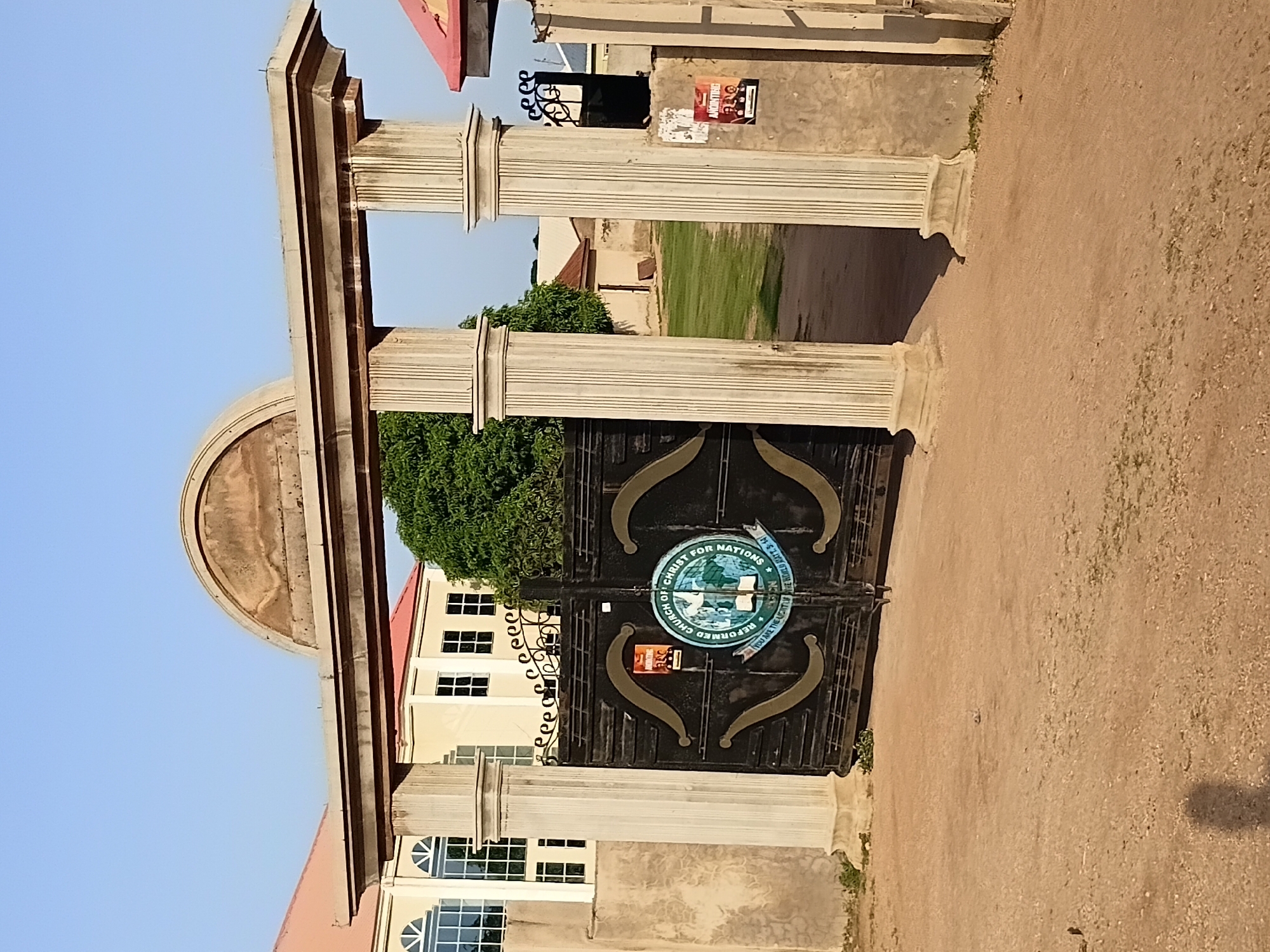 A gated entrance to a church is shown, with a large, black metal gate featuring a circular emblem and ornate scrollwork. The emblem displays a globe, a dove, and an open book, with text surrounding it that appears to read "REFORMED CHURCH OF CHRIST FOR NATIONS" and "YOU ARE THE LIGHT OF THE WORLD MATT. 5:14". Two posters are attached to the gate's right pillar; one displays the word "ANNOUNCING" above a photo of three people, and the other is red with white text. The gate is set within a concrete archway supported by two fluted pillars on either side. Behind the gate, a grassy area is visible, with buildings and trees in the background. To the left of the gate, a light-colored building with large windows is partially visible. The ground in front of the gate is dirt and gravel. The scene is set in Jalingo, Nigeria.