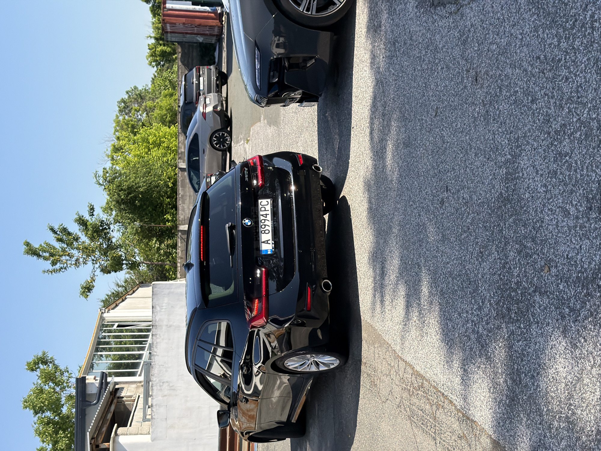 The image captures an outdoor parking area on a clear, sunny day, with the view rotated 90 degrees clockwise. The main subject is a black BMW station wagon (likely a 3 Series Touring) parked on asphalt, seen from its rear. Its taillights are visible, along with dual exhaust tips. The prominently displayed Bulgarian license plate reads "A 8994 PC," with "BG" below, indicating the car is registered in Bulgaria, specifically the Burgas region ('A' is the regional code for Burgas).
Behind the black BMW, a line of other parked vehicles is visible, including a lighter-colored hatchback and several darker cars. To the right of the BMW, the front section and wheel of another dark, modern car are visible, suggesting parallel parking.
The ground is a dark grey, textured asphalt, showing some cracks. Strong, defined shadows from the cars and surrounding structures fall on the pavement, indicative of bright sunlight during midday or early afternoon. In the background, there are various buildings, some appearing industrial with metal containers, and others with windows and glass facades. Lush green trees are visible against the clear blue sky, bordering the parking area. No people are present in the scene, which depicts a static moment of parked vehicles.
