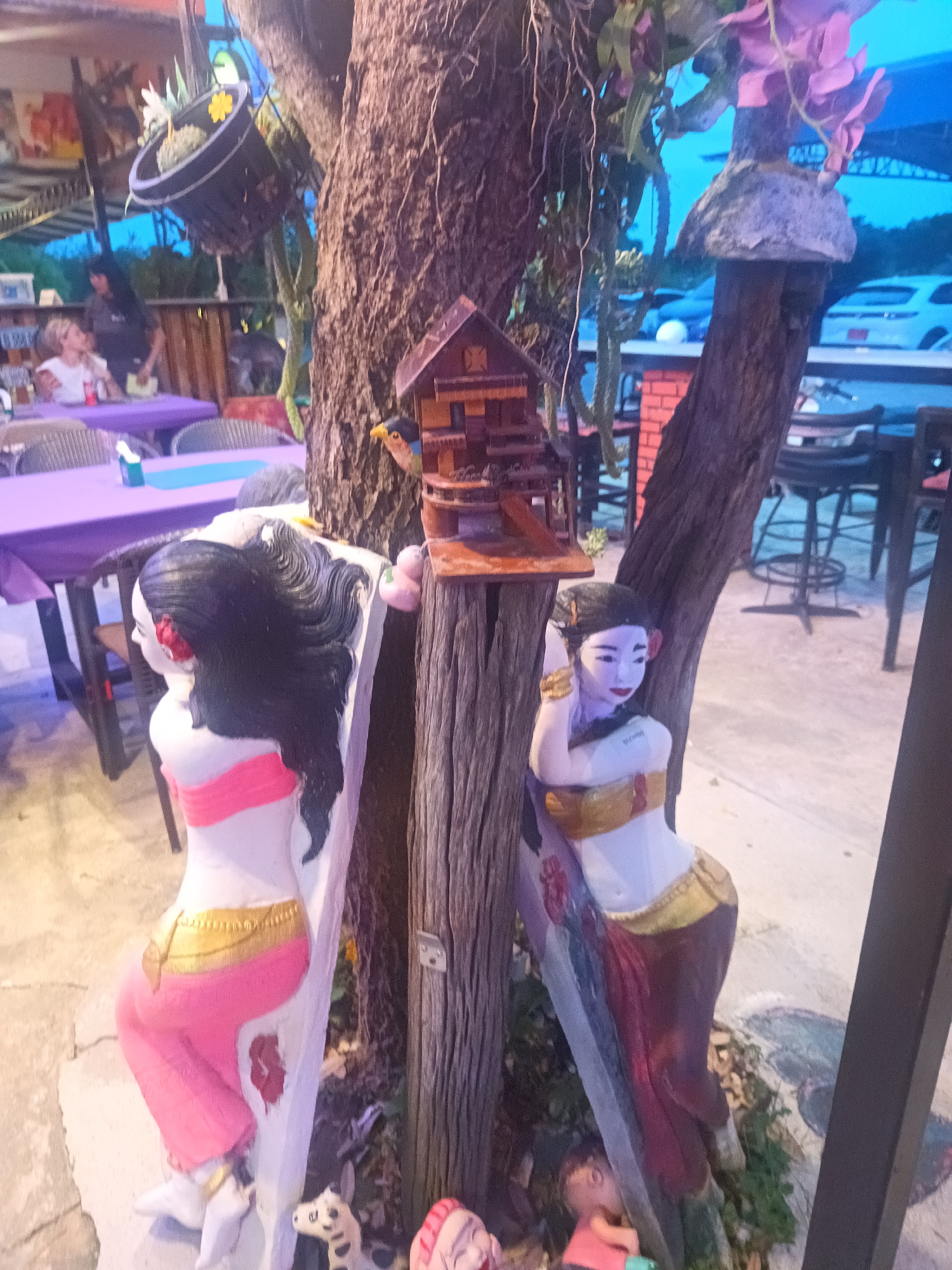 In Ban Nong Khon, Thailand, the image captures a scene at an outdoor establishment. Two painted statues of women flank a weathered tree trunk. A small, wooden decorative birdhouse is attached to the tree. Nearby, there are tables covered with purple cloths and chairs, occupied by at least two people. A hanging basket with yellow flowers is suspended above the scene. In the background, partially visible, are parked vehicles and the open sky. The setting appears to be a restaurant or gathering place.