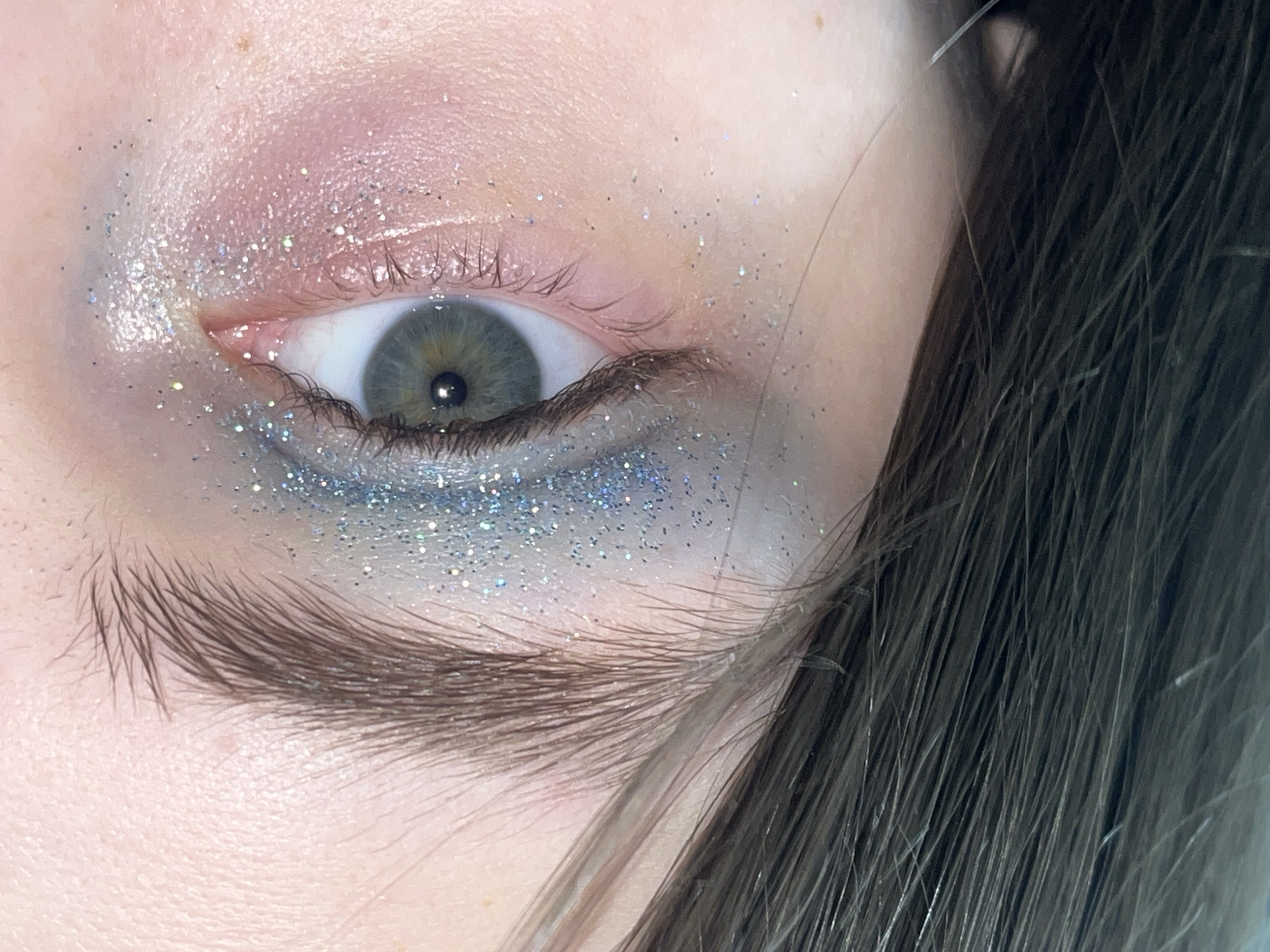 A close-up image shows a human eye with elaborate makeup. The upper eyelid features a blend of pink and iridescent eyeshadow, accented with scattered glitter. The lower eyelid is adorned with a heavier concentration of blue glitter. The eyebrow is full and dark. The iris is a mix of grey and green with a dark pupil. Skin texture is visible, including small imperfections. Strands of dark hair partially obscure the right side of the face. There is no indication of location.