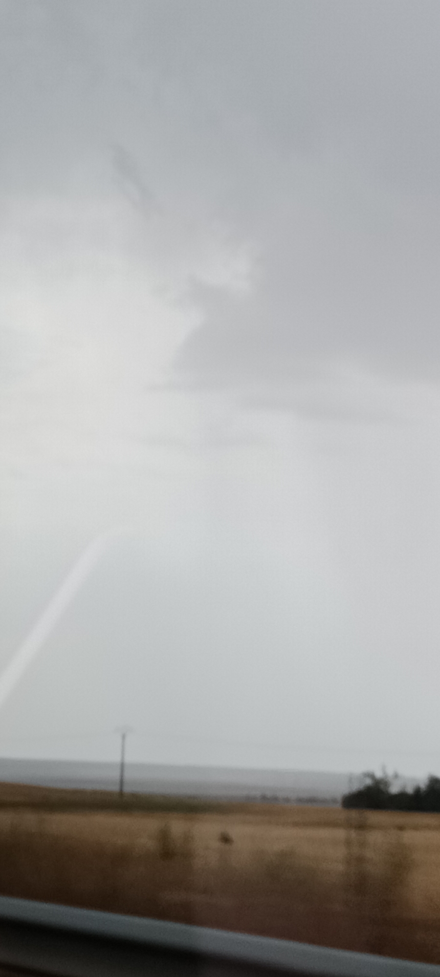 This is a vertically oriented, blurry, low-resolution, outdoor photograph taken from a moving vehicle. The image depicts a grey, overcast sky with some faint clouds and what appears to be a partial rainbow arc on the left side. Below the sky is a distant horizon with a thin line of darker land or water. In the middle ground, there is a wide, brown field with some vegetation, and a single, thin pole with crossbars, possibly a utility pole, is visible in the center of the frame. In the foreground, a dark, blurry stripe runs horizontally across the bottom of the image, suggesting it is the edge of the vehicle from which the photo was taken. The scene is drab and lacks sharp detail. It is located in the rural outskirts of Cerecinos de Campos, Spain.