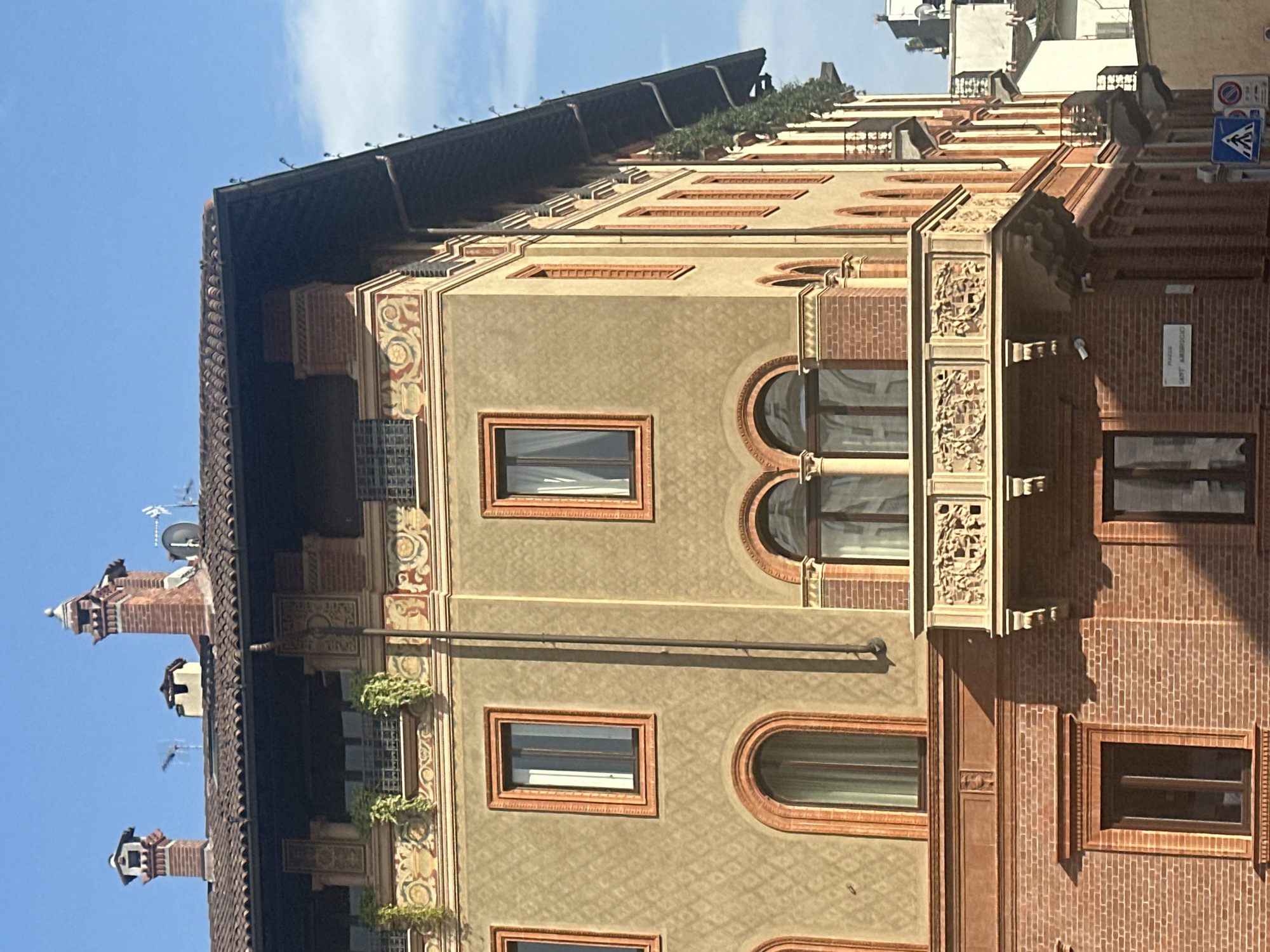 The image shows the corner of an ornate building in Milan, Italy, with a clear blue sky. The building features a terracotta-tiled roof with several decorative chimneys. The facade is a mix of stucco and exposed brick, with intricate decorative friezes and patterned stucco work. There are several arched windows with reddish-orange frames, and a prominent balcony with elaborate carved details. Greenery spills over the edges of balconies on higher floors. A street sign that reads "PIAZZA SANT'AMBROGIO" is visible on the lower part of the building. Traffic signs and a pedestrian crossing are visible on the sidewalk. The overall impression is of historic and well-maintained architecture.