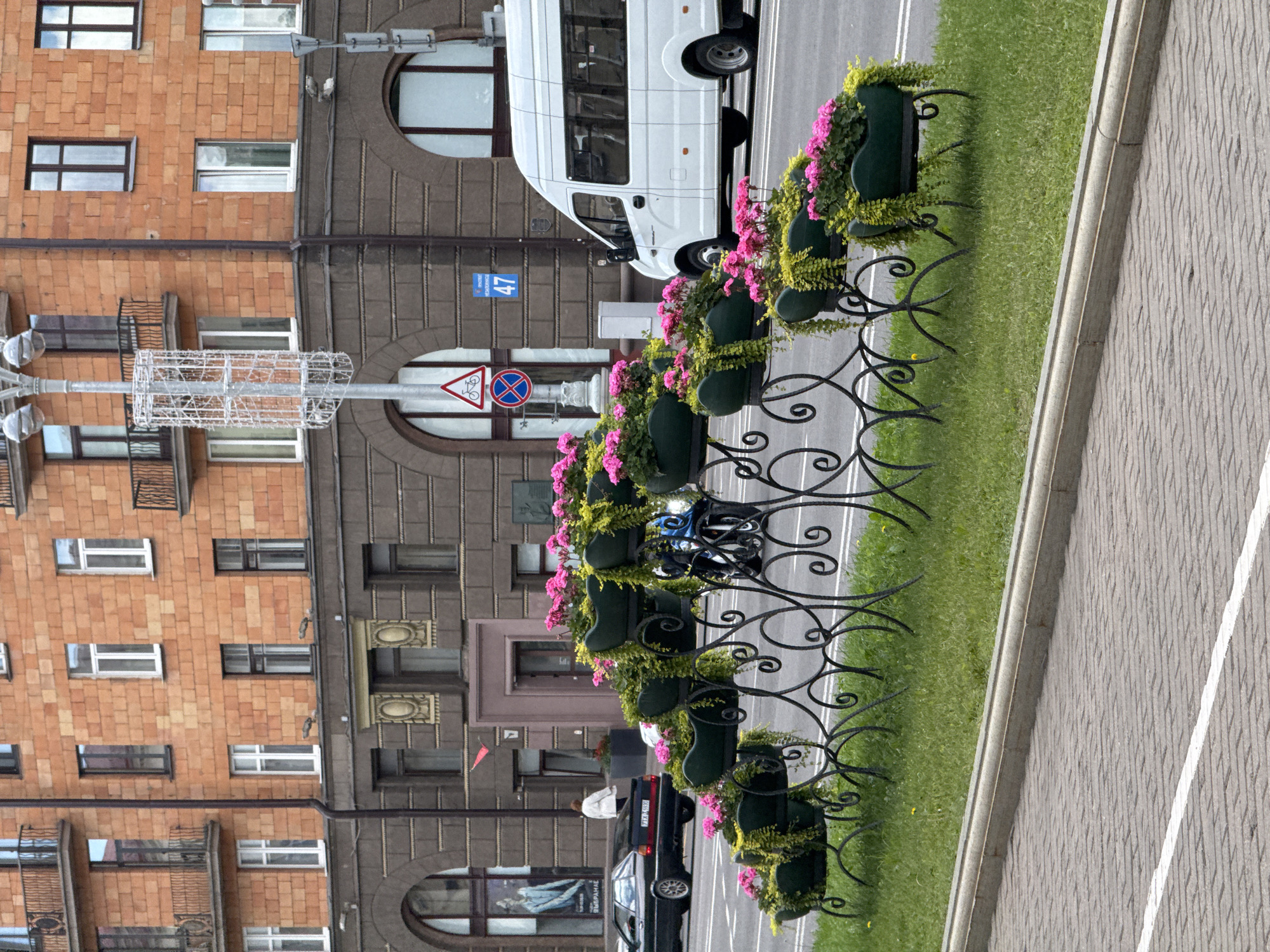 An outdoor daytime scene on a street in Minsk, Belarus, shows a tiered arrangement of green planters filled with pink flowers and trailing greenery, set on a decorative black metal stand. The planters are positioned on a grassy verge separating the sidewalk from the roadway. Behind the planters, a white mini-van is parked to the right, and a dark-colored sedan is visible on the road to the left. In the background, a multi-story brick building with arched ground-floor windows and balconies on upper floors dominates the scene. A lamppost with decorative cages stands near the building's corner. A blue street sign with the number "47" is mounted on the building. The sidewalk in the foreground is paved with grey brickwork and has a white line marking.