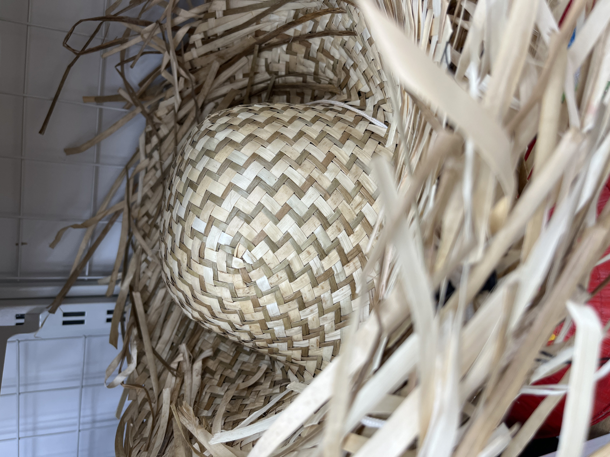 The image shows a woven basket inside a straw hat, both appearing to be made from natural fibers. The basket has a rounded shape and is woven in a pattern of alternating light and dark colors. The straw hat surrounds the basket with a chaotic array of straw pieces. The background shows a portion of a white wire shelf and possibly some red or dark-colored items partially obscured by the straw. The location is inferred to be Bac Nha Trang Ward, Vietnam, but there are no specific elements in the image to confirm this.
