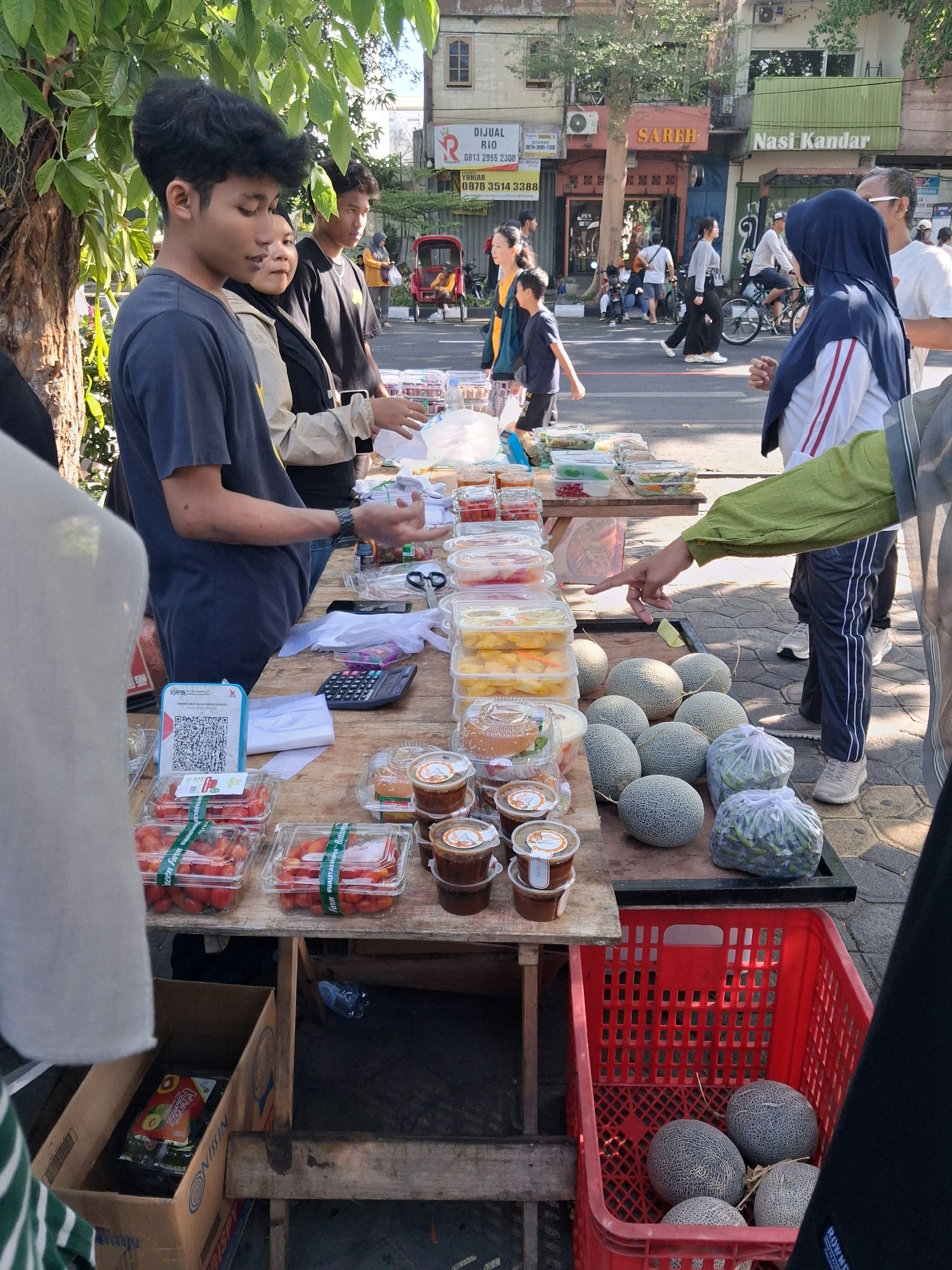 A street vendor is selling produce from a wooden stall on the side of a street in Surakarta, Indonesia. A young man in a dark blue t-shirt is attending to the stall, interacting with customers. Nearby, other people are walking by, some on bicycles. The stall is laden with various food items, including containers of tomatoes, fruit, and sauces, as well as several cantaloupes. A red plastic crate filled with cantaloupes sits in the foreground. The background shows a street with buildings, signs, and trees.