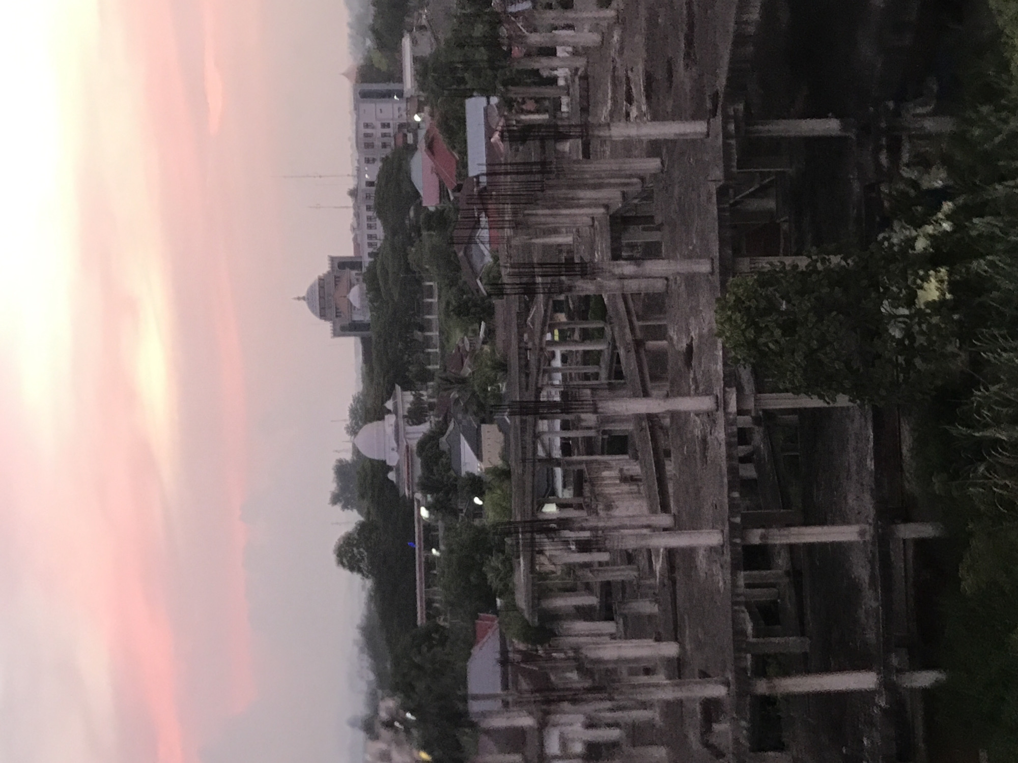 The image, oriented horizontally, depicts an urban landscape in Cotbadatunong, Indonesia, bathed in the soft light of either sunrise or sunset. The upper half of the frame is dominated by a hazy sky, exhibiting a gentle gradient of light pink and pale orange, fading into a brighter, lighter hue towards the left.
In the foreground, a dense, skeletal structure of concrete pillars and exposed rebar signifies an unfinished or abandoned construction project. This unfinished edifice creates a stark, angular contrast against the softer background. Beyond the construction site, the middle and background layers showcase a scattering of green trees and various buildings. Several structures feature distinct domes, indicative of mosques or other significant public architecture common in the region. The overall lighting is subdued, suggesting a peaceful, transitional moment of day. No people or discernible text are visible in the scene.